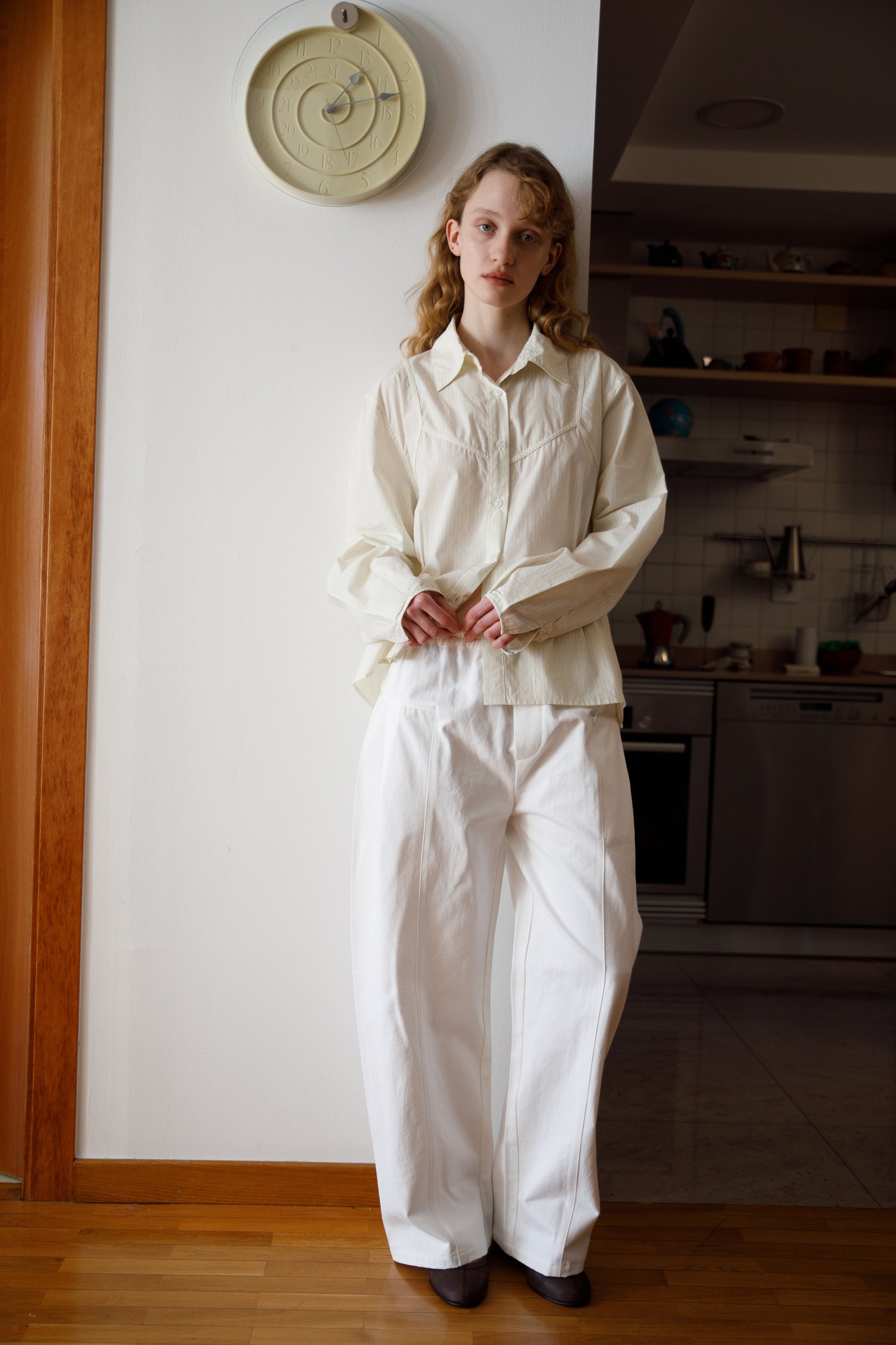A person with long, wavy blonde hair stands indoors by a white wall, wearing a loose-fitting cream shirt and Pleat Pant - White. A clock hangs above them, with a kitchen visible in the background.