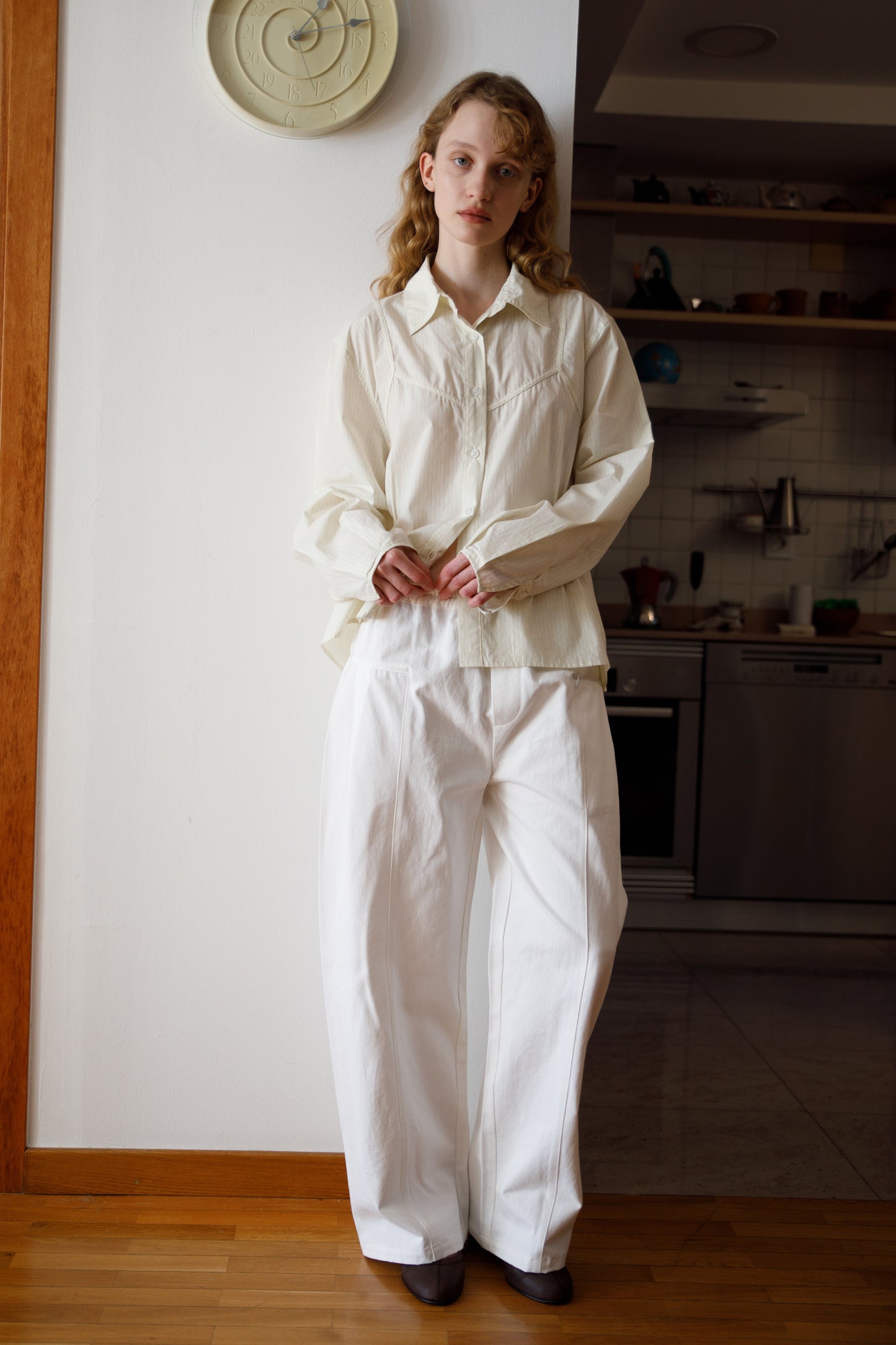 A woman with light hair stands indoors by a white wall, wearing the Cami Overshirt - Citrus Stripe. She is on a wooden floor near a kitchen area, with a clock on the wall above her.