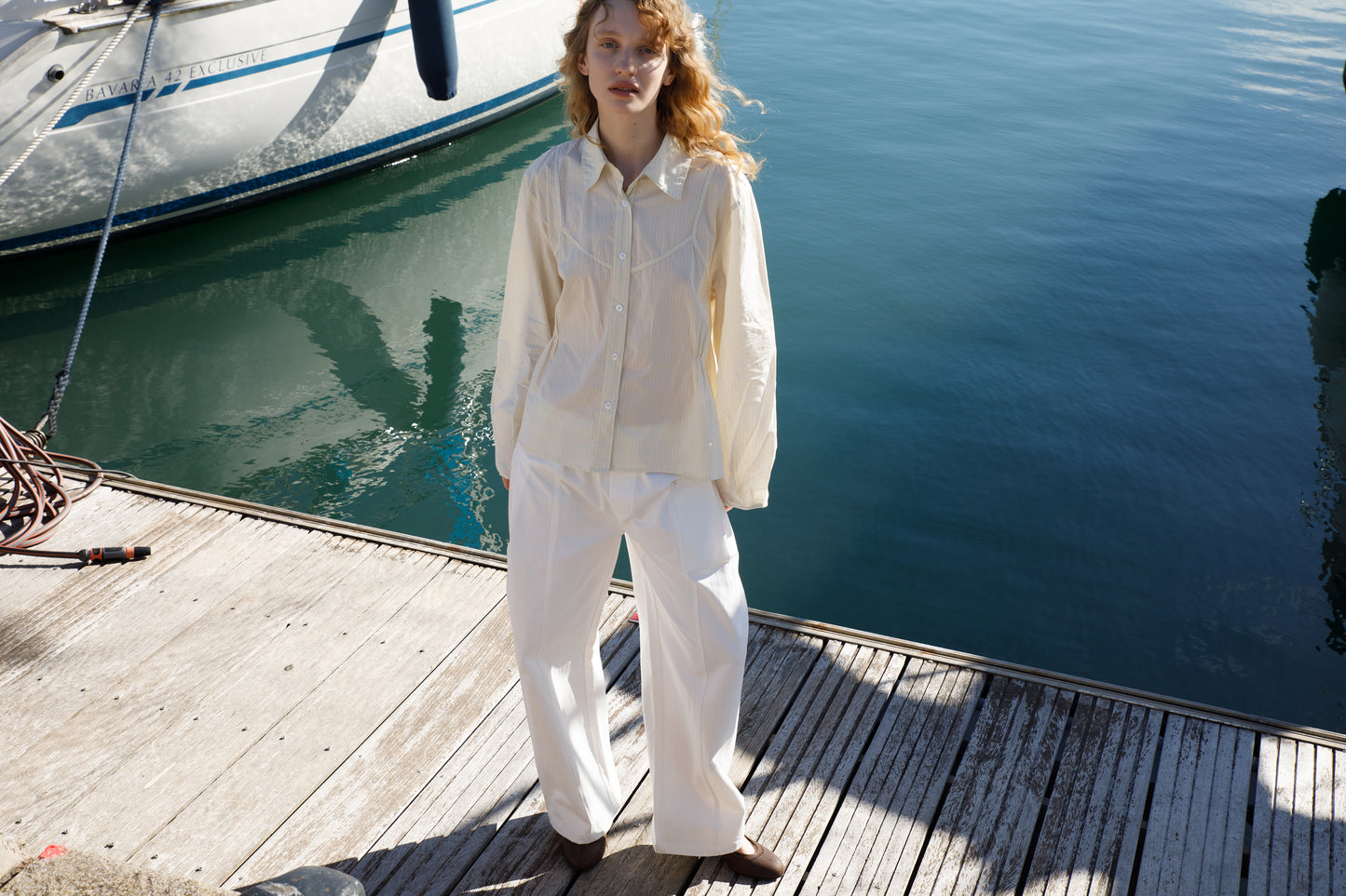 A person with wavy blond hair stands on a dock near a sailboat, wearing the Cami Overshirt - Citrus Stripe from Deiji Studios and white wide-legged pants, with calm blue water in the background.