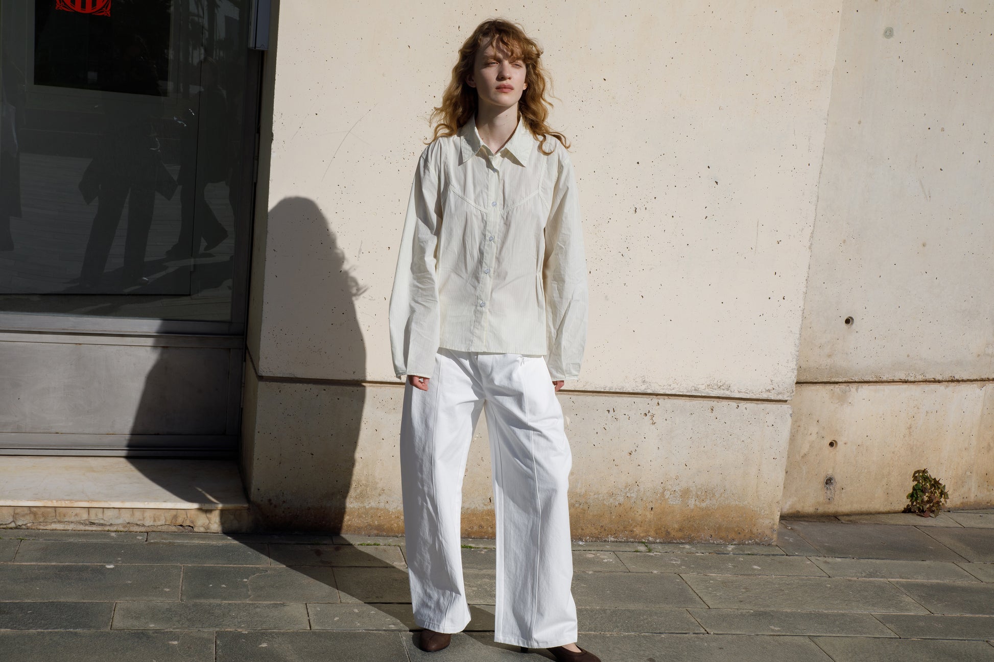 A person with wavy hair wears a loose-fitting white shirt and the Pleat Pant - White, standing on a sunlit sidewalk by a beige wall. Their hands rest in their pockets as they look ahead with a neutral expression.