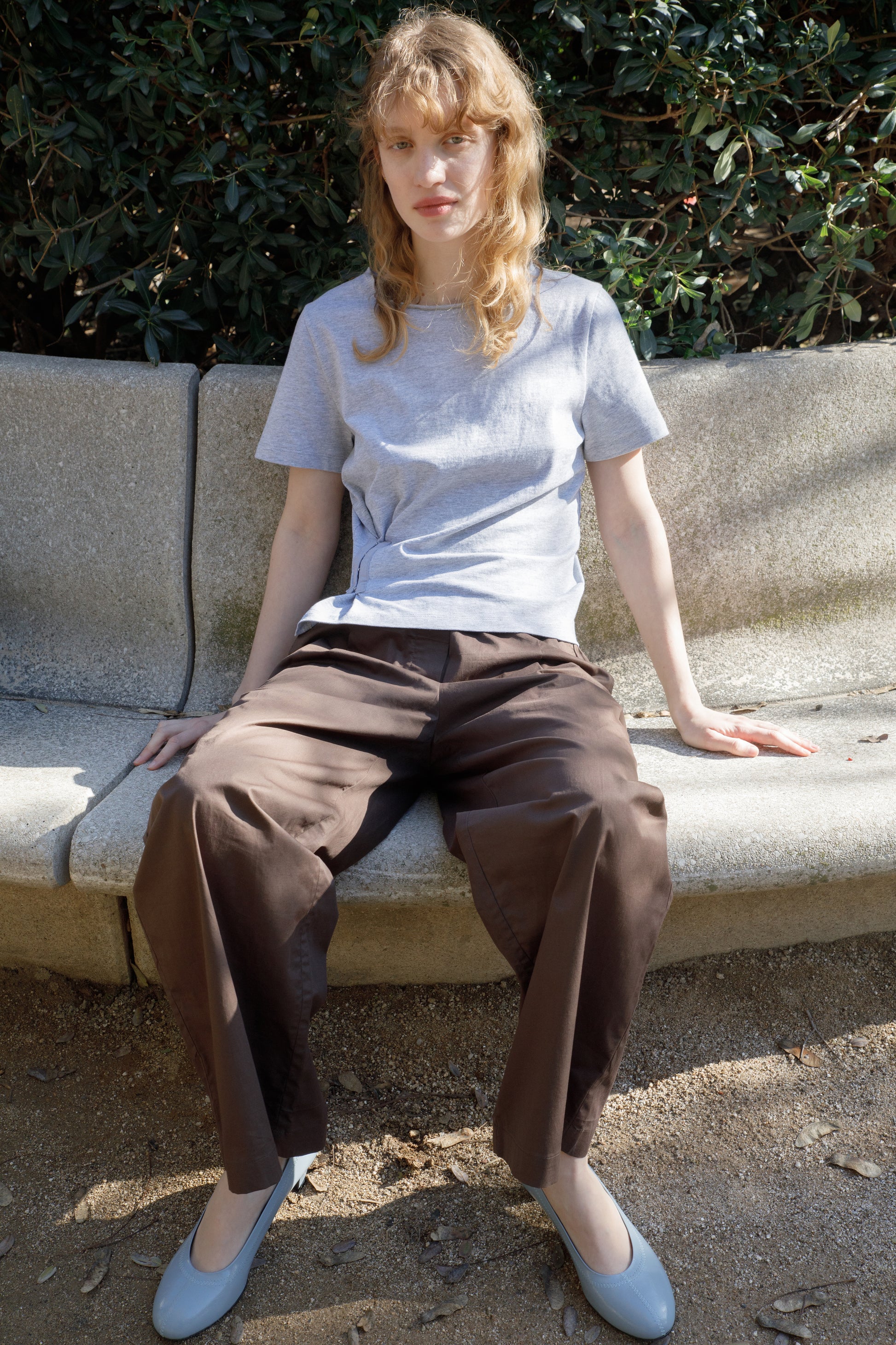 A woman with wavy blonde hair sits on a curved stone bench outdoors, wearing a light gray T-shirt and the Contrast Waist Pant - Brown. She looks directly at the camera, with greenery in the background.