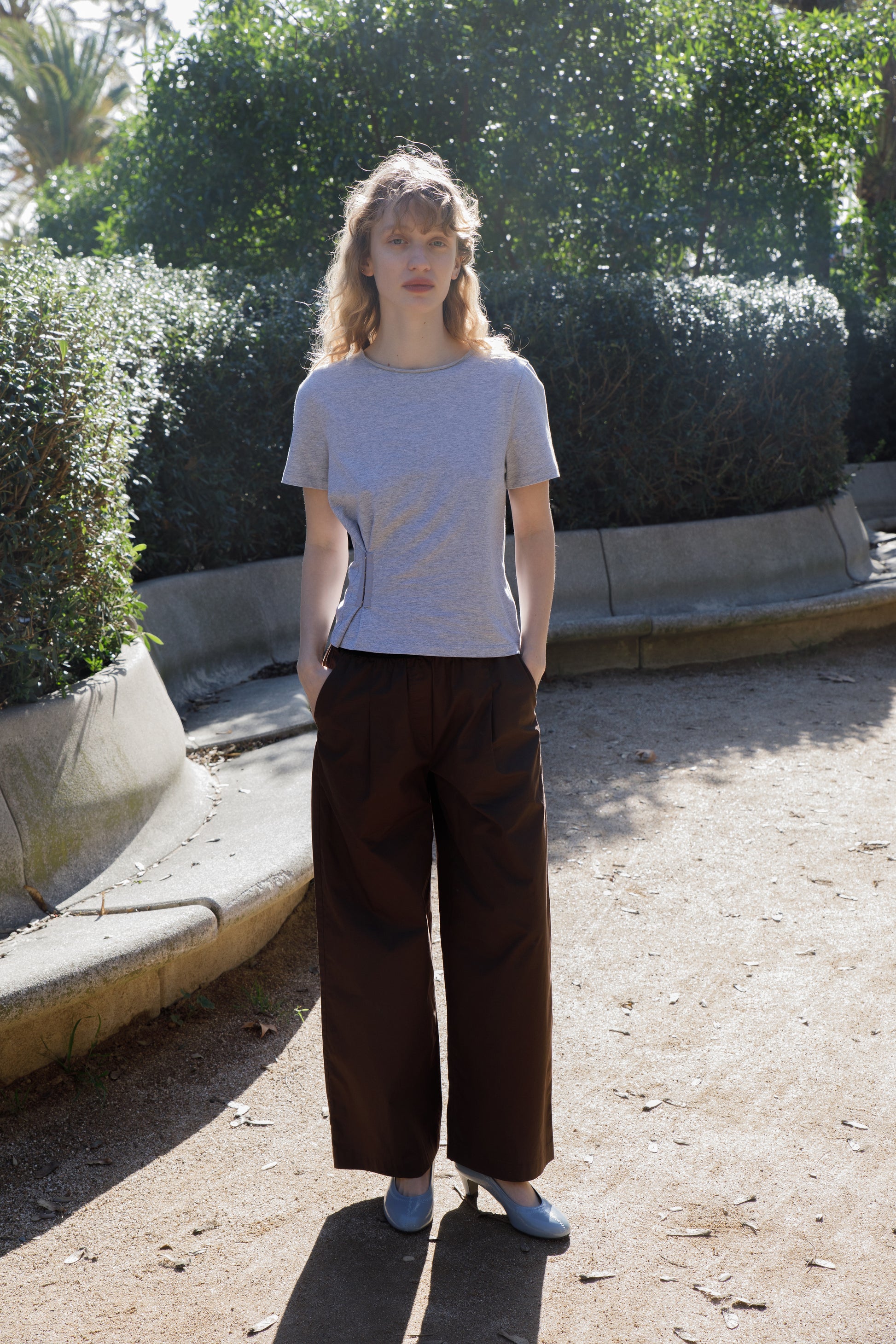A person with curly blond hair stands on a sunlit park pathway, wearing a gray t-shirt, Contrast Waist Pant - Brown, and light blue shoes, surrounded by green bushes.