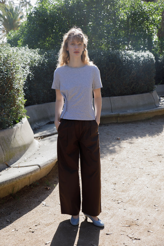 A woman with curly hair stands outdoors on a sunlit path, wearing the Pinch Tee - Grey with wide-legged dark brown pants and light blue shoes, surrounded by green bushes and concrete benches.