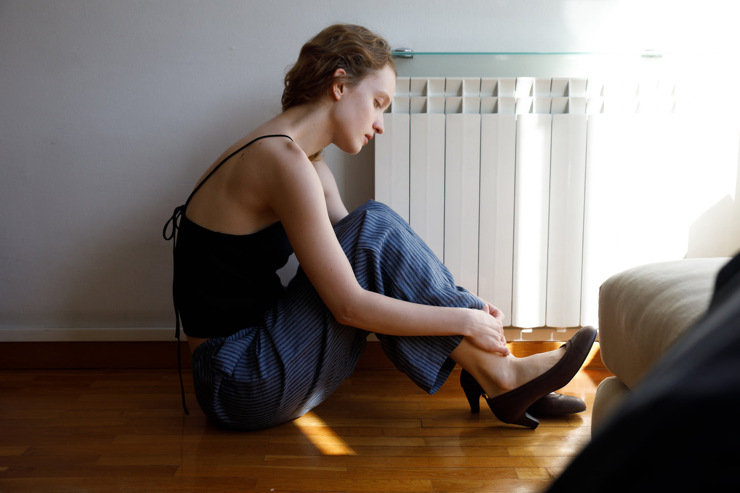 A woman sits on a wooden floor by a radiator, wearing the Linen Ease Trouser - Azure Stripe from Deiji Studios and a black sleeveless top. She adjusts her black heels as sunlight streams in from the right.