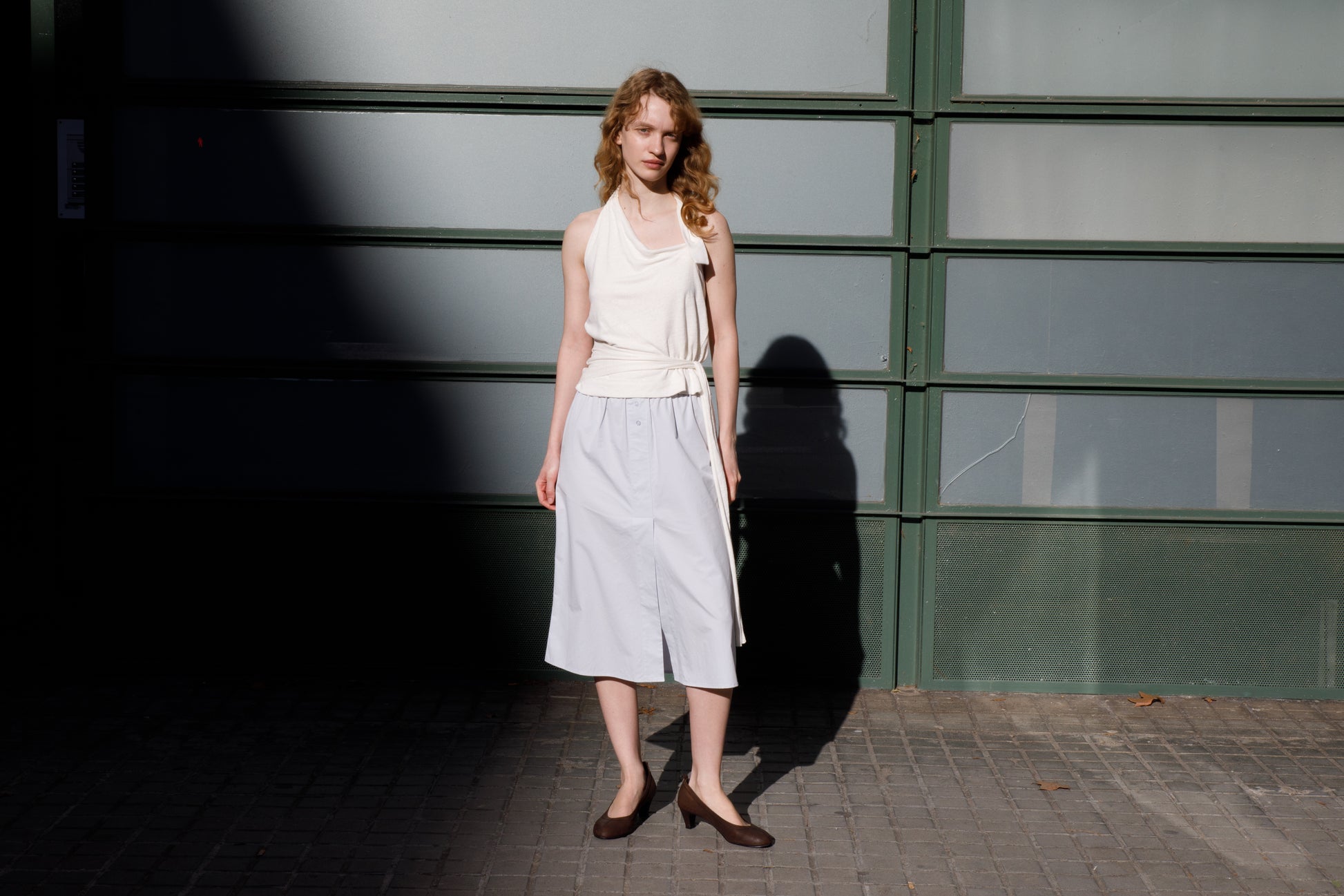 A woman with curly hair stands on a sidewalk by a green and frosted glass wall, wearing a sleeveless white top, the Overlap Skirt - Blue from Deiji Studios, and dark flats as sunlight and shadows fall across the scene.