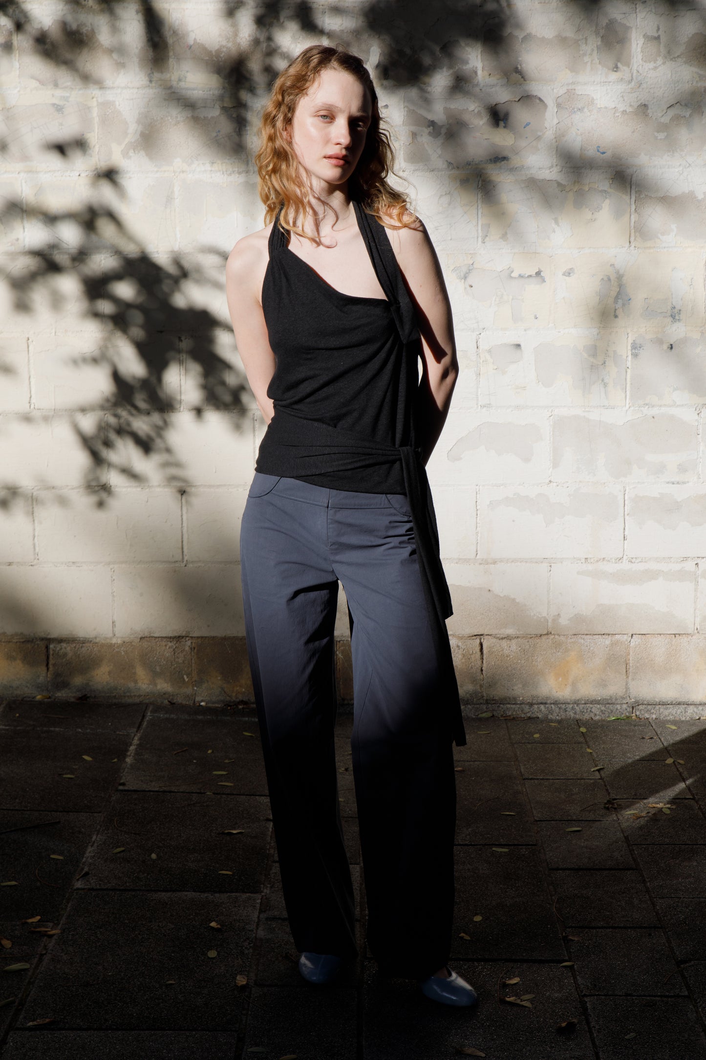A person with wavy, shoulder-length hair stands before a sunlit white brick wall, wearing a sleeveless black top and the Panelled Pant - Charcoal by Deiji Studios as tree-branch shadows dance across the scene.