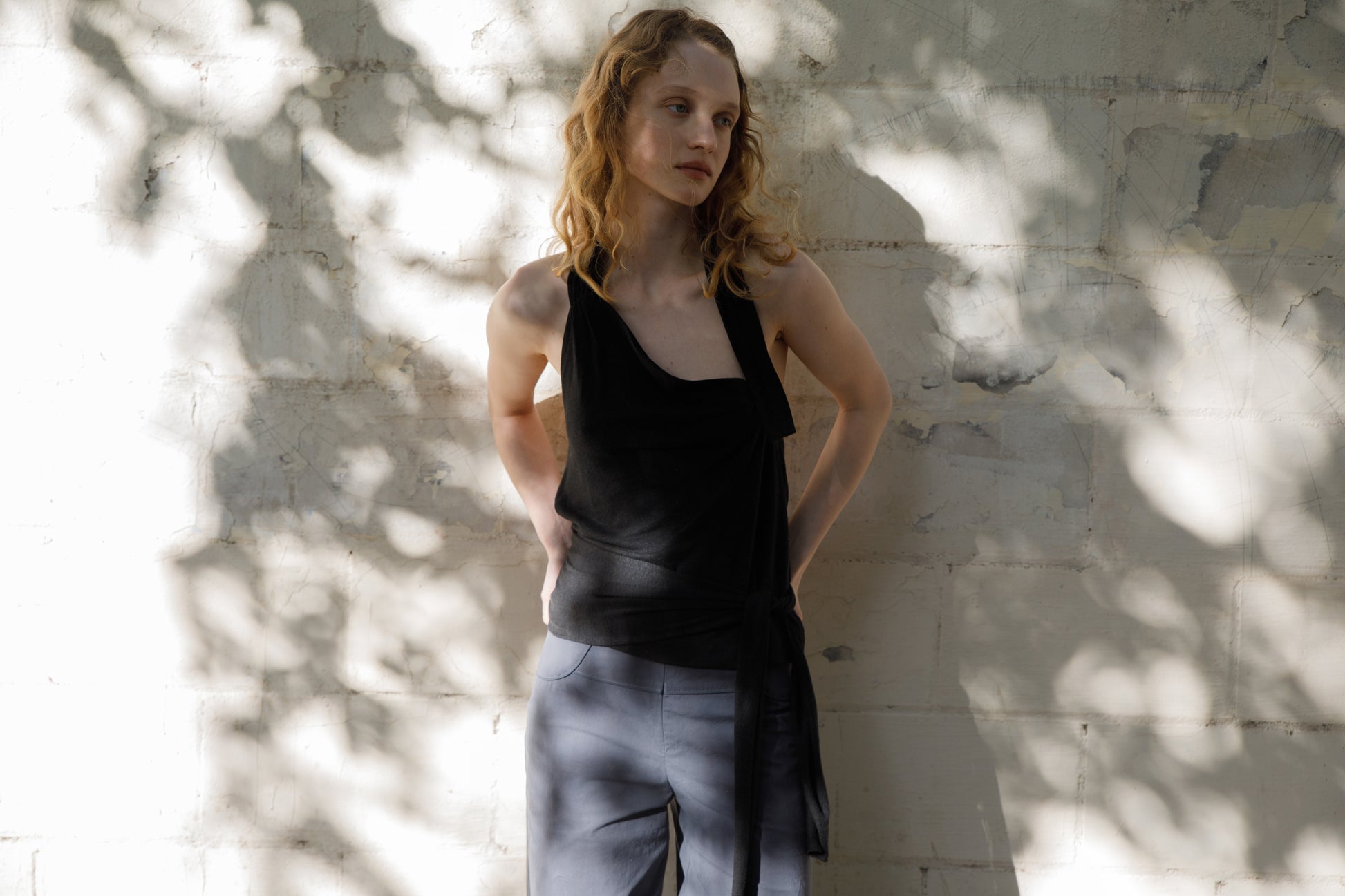 A woman with wavy blonde hair stands against a sunlit, textured wall, wearing a black sleeveless top and the Panelled Pant - Charcoal in a straight-leg silhouette. Dappled shadows of leaves play across the wall behind her.