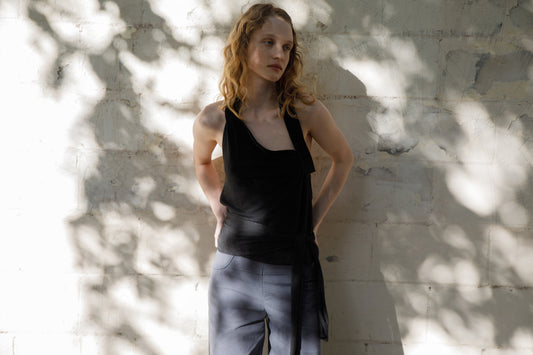 A woman with wavy blonde hair stands against a sunlit, textured wall, wearing a black sleeveless top and the Panelled Pant - Charcoal in a straight-leg silhouette. Dappled shadows of leaves play across the wall behind her.