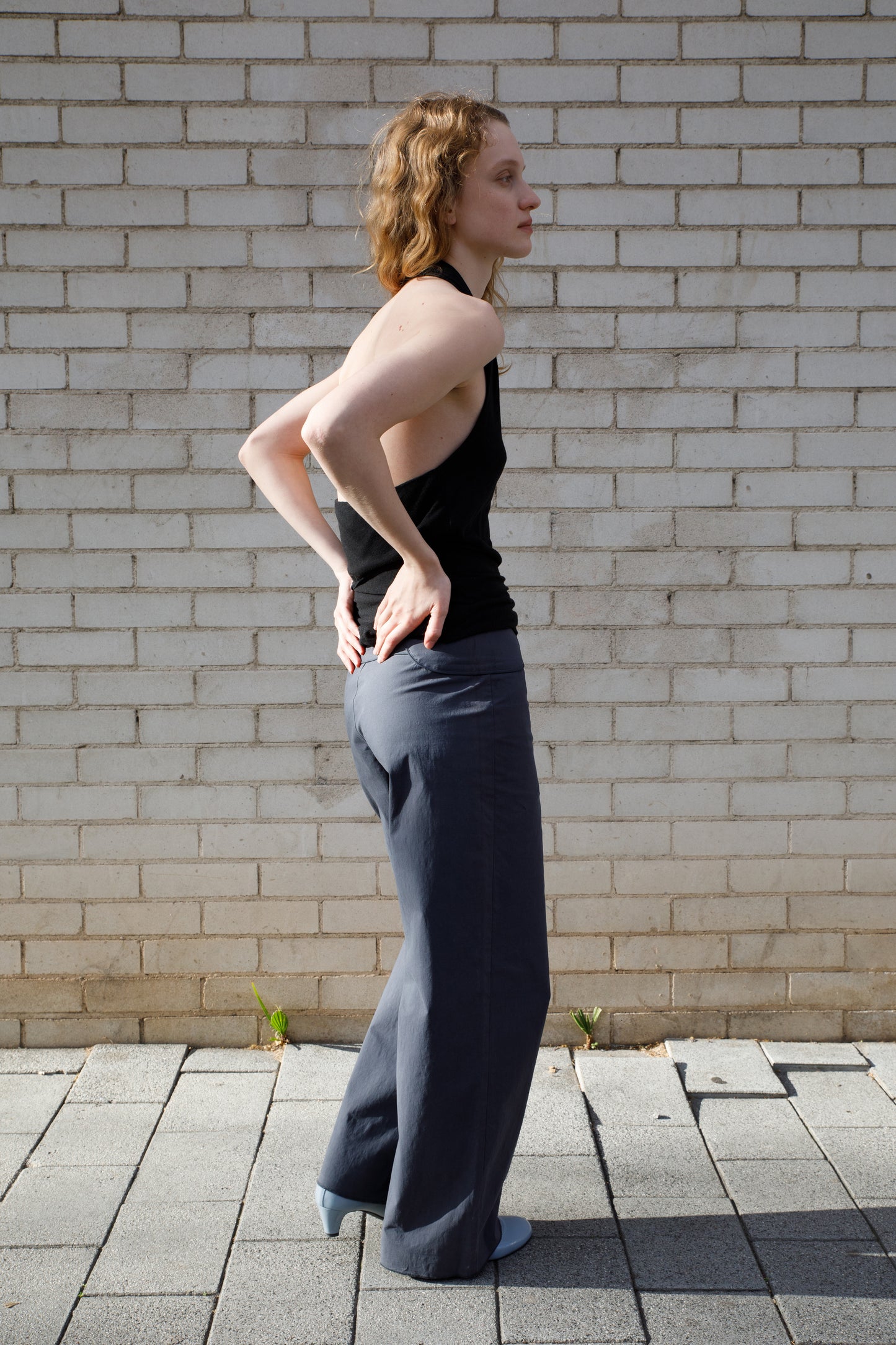 A person with shoulder-length wavy hair stands sideways against a white brick wall, wearing a black sleeveless top, Deiji Studios Panelled Pant - Charcoal in a straight-leg style, and light blue shoes, with hands on their lower back.