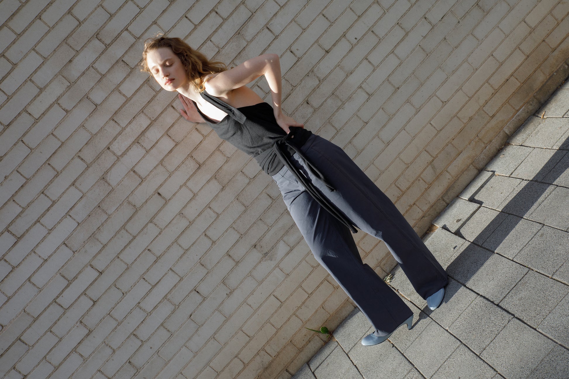 A woman in a black tank top and Deiji Studios Panelled Pant - Charcoal with a straight-leg silhouette poses against a white brick wall on a sunny day, her blue shoes and shadow adding contrast.
