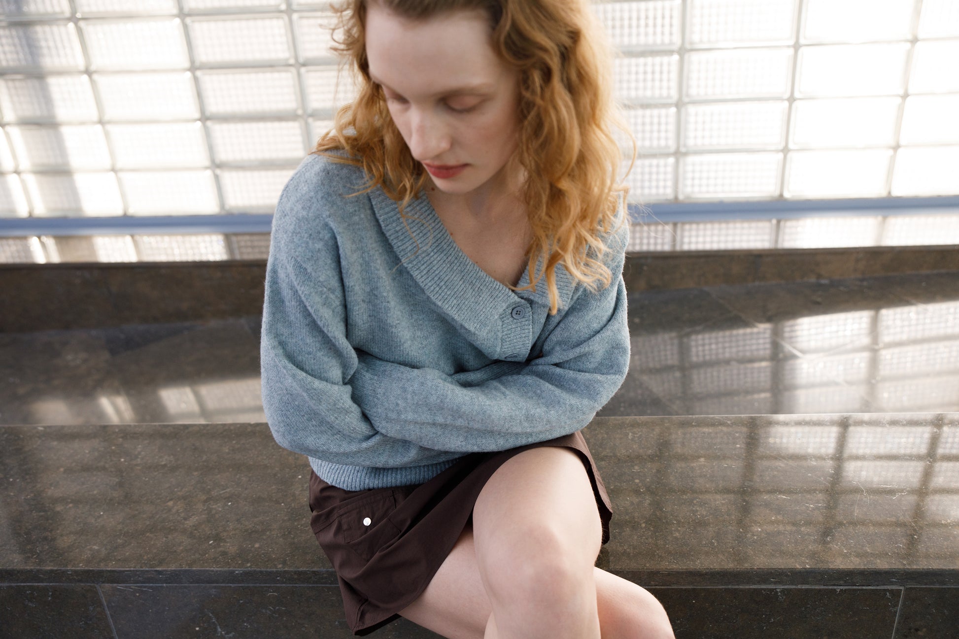 A woman with wavy, light brown hair sits on a stone bench with her arms crossed, looking down. She wears a loose gray sweater and the Pocket Skirt in Sepia, set against a glass block wall reflecting Deiji Studios’ minimalist style.