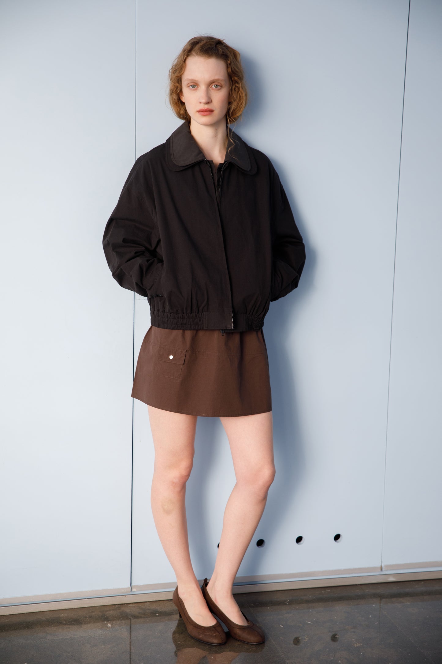 A model wears the Collared Zip Jacket - Black Contrast by Deiji Studios, styled with a brown mini skirt and ballet flats, standing against a light blue wall.