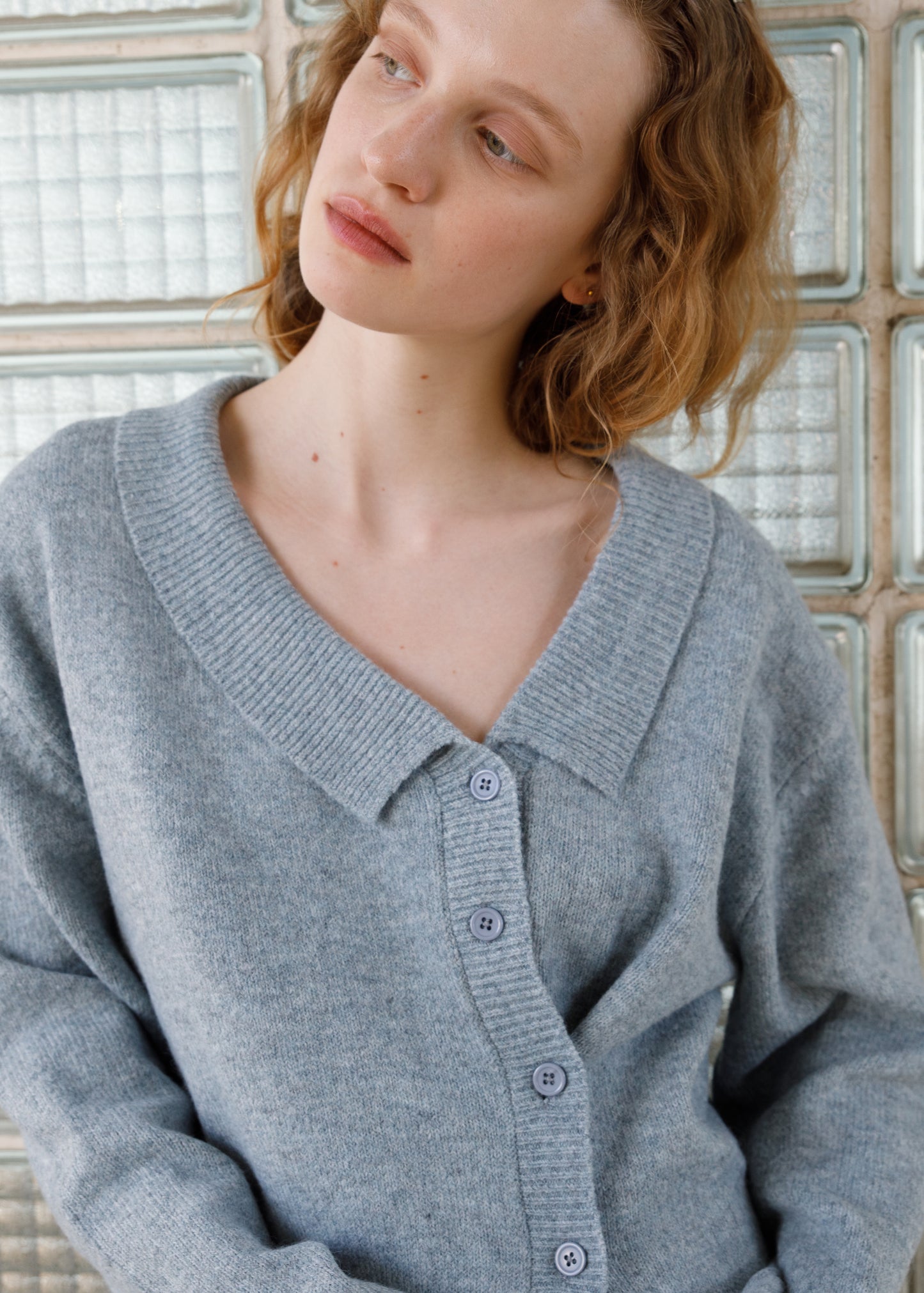 A woman with wavy, shoulder-length hair in an Ice Blue Cross Button Cardigan gazes thoughtfully to the side, standing before a wall of glass blocks.