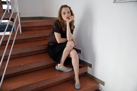 A young woman with long, wavy hair sits thoughtfully on a red staircase indoors, wearing the Deiji Studios Close Shirt in Sepia and light blue shoes.