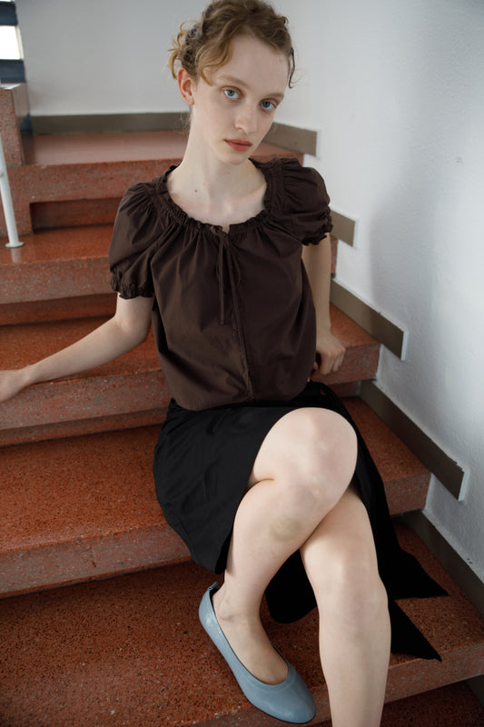 A young woman with light skin and curly hair sits on red stairs, wearing the Close Shirt - Sepia with puffed sleeves, a black skirt, and gray flats, looking directly at the camera with a neutral expression.