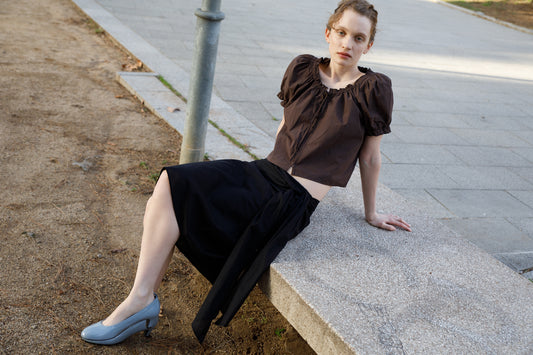 A woman with light skin and brown hair, dressed in a brown puff-sleeve top, black Scarf Midi skirt by Deiji Studios, and blue heels, sits outdoors on a stone bench with one arm resting back and one leg extended.