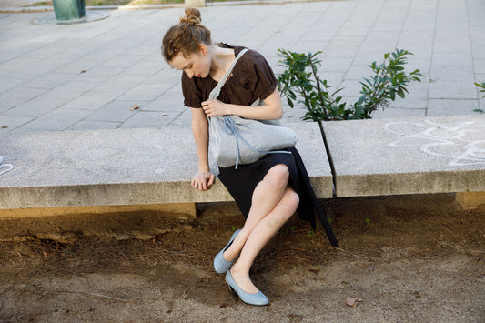 A woman with light hair tied up sits on a concrete bench outdoors, wearing Deiji Studios' Close Shirt - Sepia, a black skirt, and light blue flats. She looks down at the gray handbag on her lap, with shrubs visible in the background.