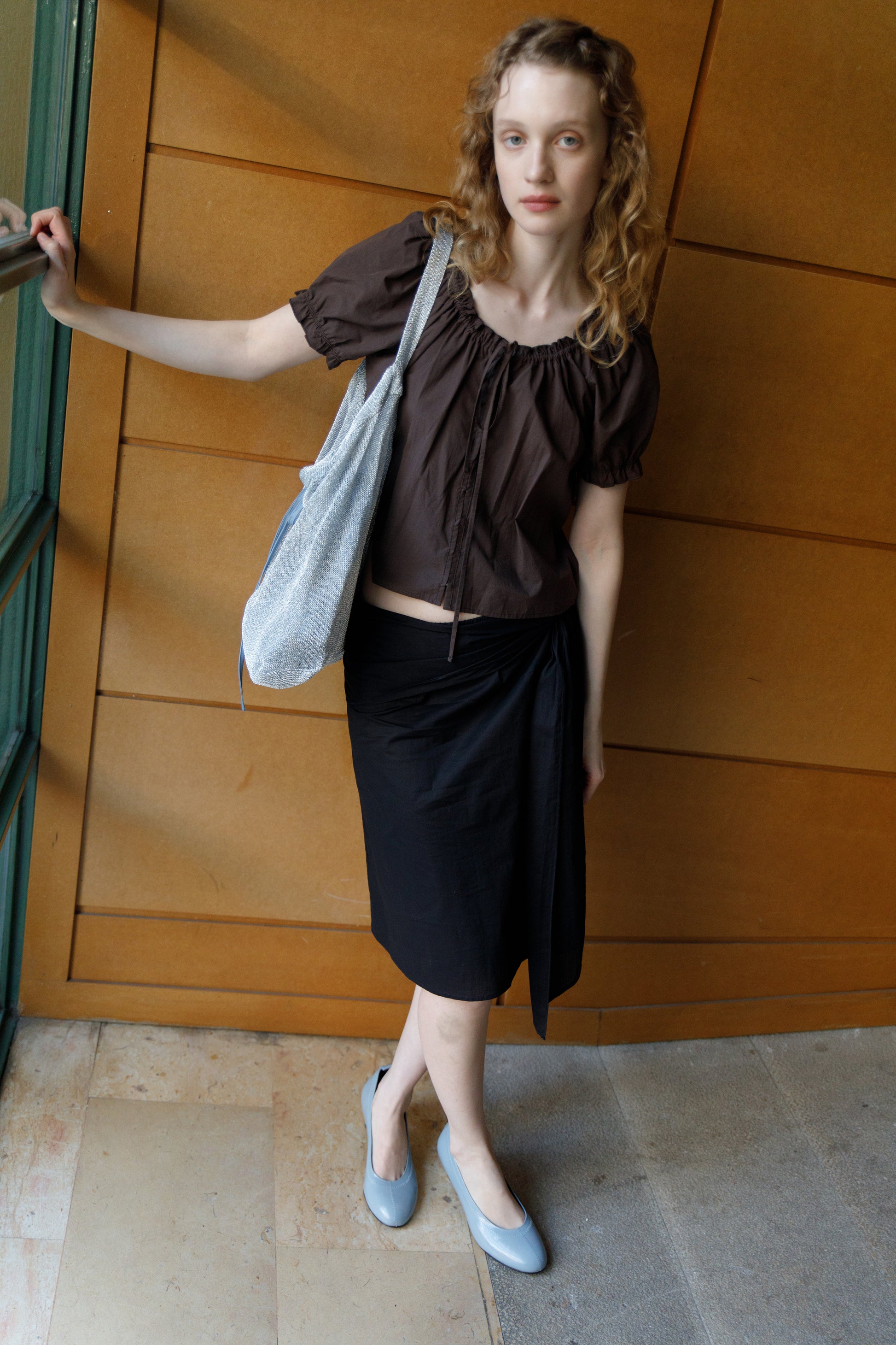 A woman with wavy blonde hair stands indoors by a wooden wall, wearing a Deiji Studios Close Shirt - Sepia with puffed sleeves, a black wrap skirt, gray flats, and holding a light-colored shoulder bag.