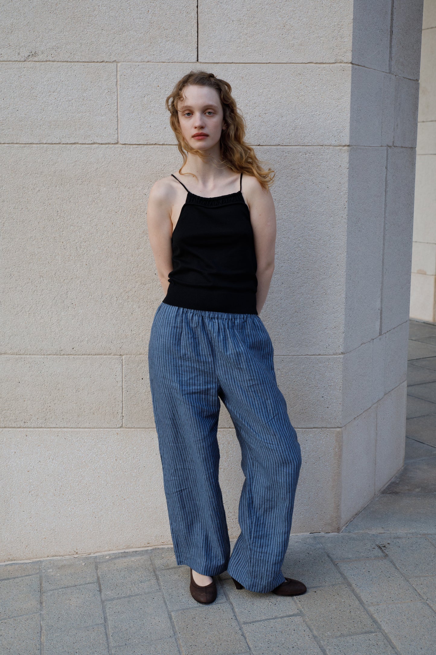 A woman with wavy blonde hair stands against a light stone wall, wearing a black sleeveless top and the Linen Ease Trouser - Azure Stripe by Deiji Studios. Her hands are behind her back as she gazes at the camera.