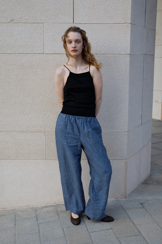 A woman with wavy blonde hair stands against a light stone wall, wearing a black sleeveless top and the Linen Ease Trouser - Azure Stripe by Deiji Studios. Her hands are behind her back as she gazes at the camera.