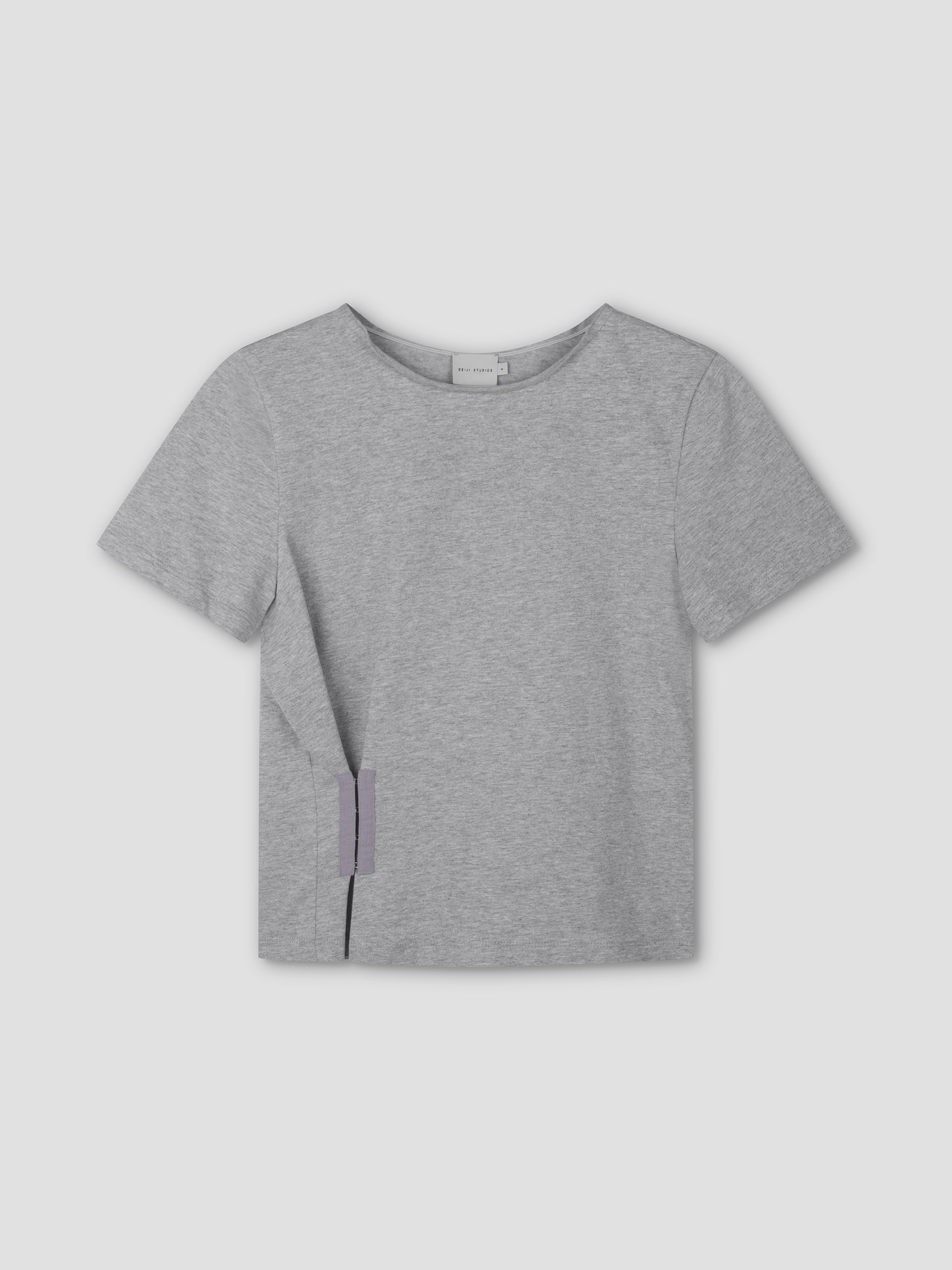The Pinch Tee - Grey is a relaxed, light gray short sleeve shirt with a round neckline, shown flat on a matching background. A small lavender tag near the left hem highlights Deiji Studios’ minimalist style.