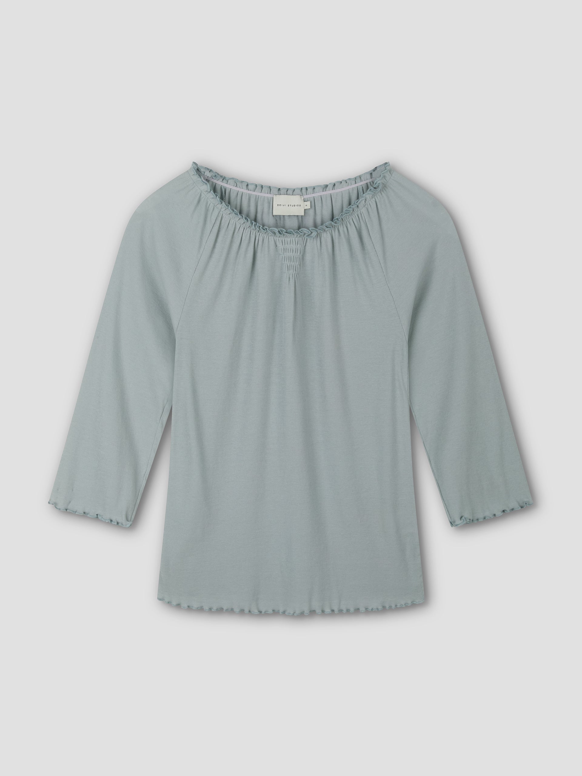 The Cotton Flounce 3/4 Sleeve - Baby Blue by Deiji Studios is a women’s top with a gathered, elastic neckline and softly ruffled edges on the sleeves and hem, shown laid flat on a plain white background.
