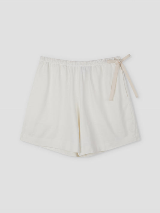 The Relax Short - Off White by Deiji Studios features a relaxed fit in white cotton with an elastic waistband and a beige ribbon bow on the right side, shown against a plain light background.