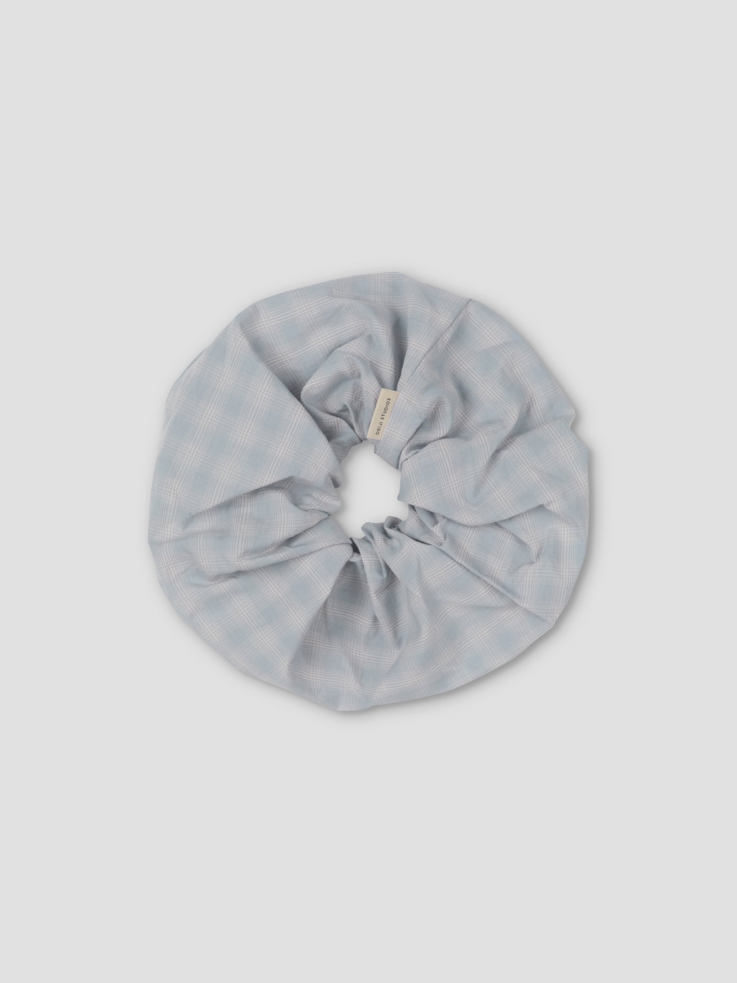 The Scrunchie is a large, light blue hair accessory with a subtle checkered pattern, shown on a plain light gray background.