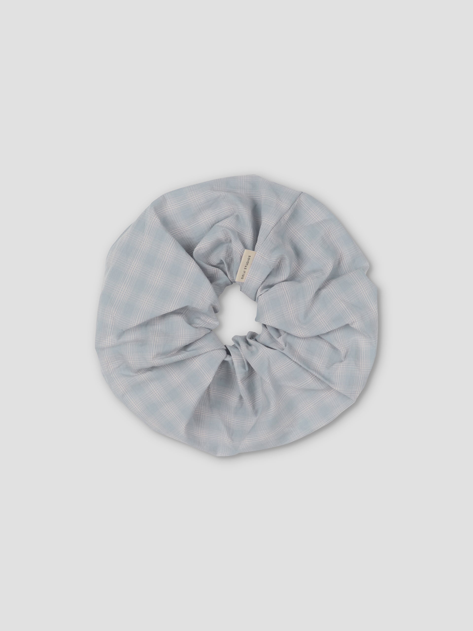 The Scrunchie is a large, light blue hair accessory with a subtle checkered pattern, shown on a plain light gray background.