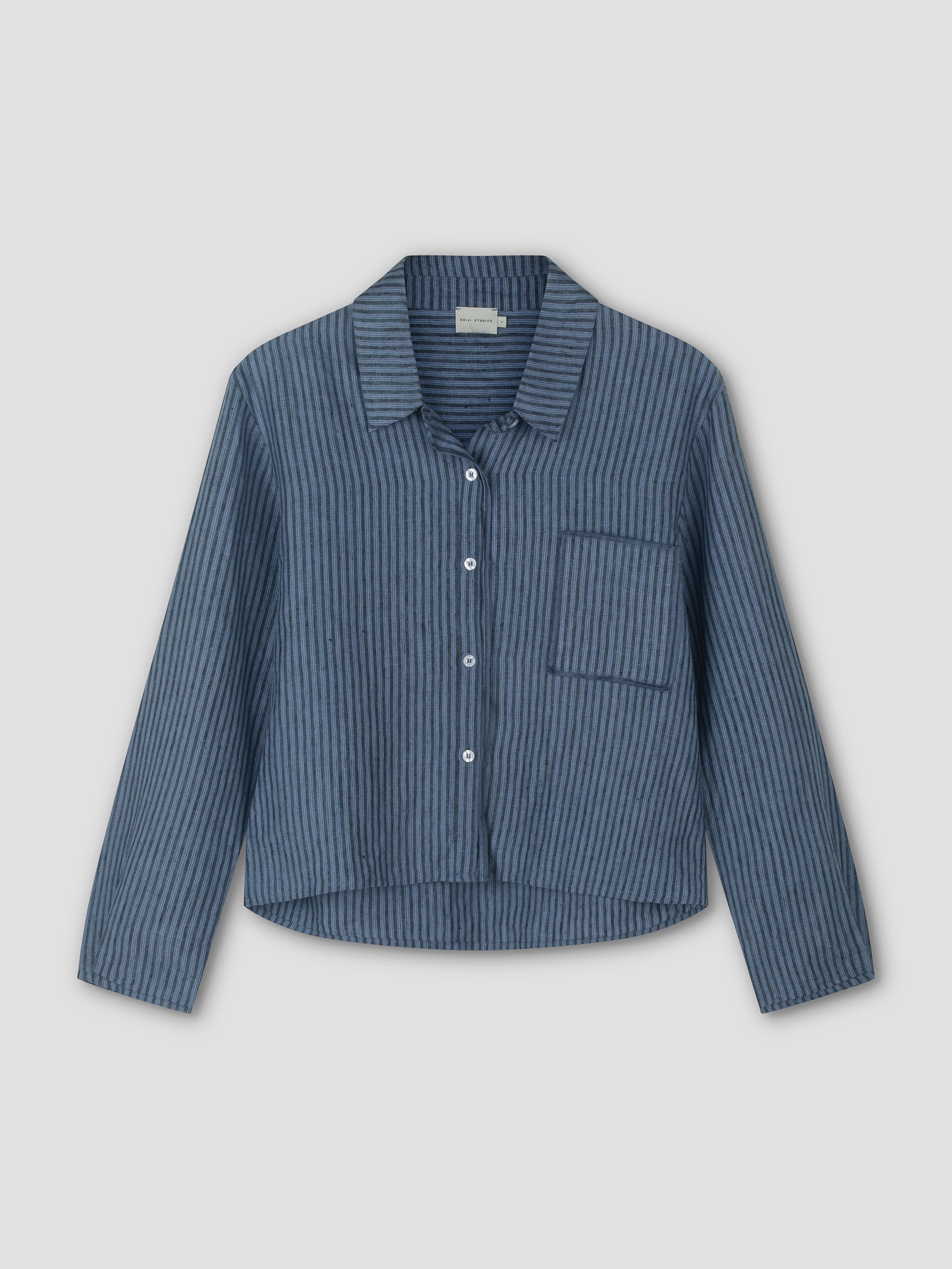 The Double Placket Shirt - Azure Stripe by Deiji Studios is a relaxed long sleeve with blue and dark blue vertical stripes, left chest pocket, collar, and button-up front, shown against a light gray background.