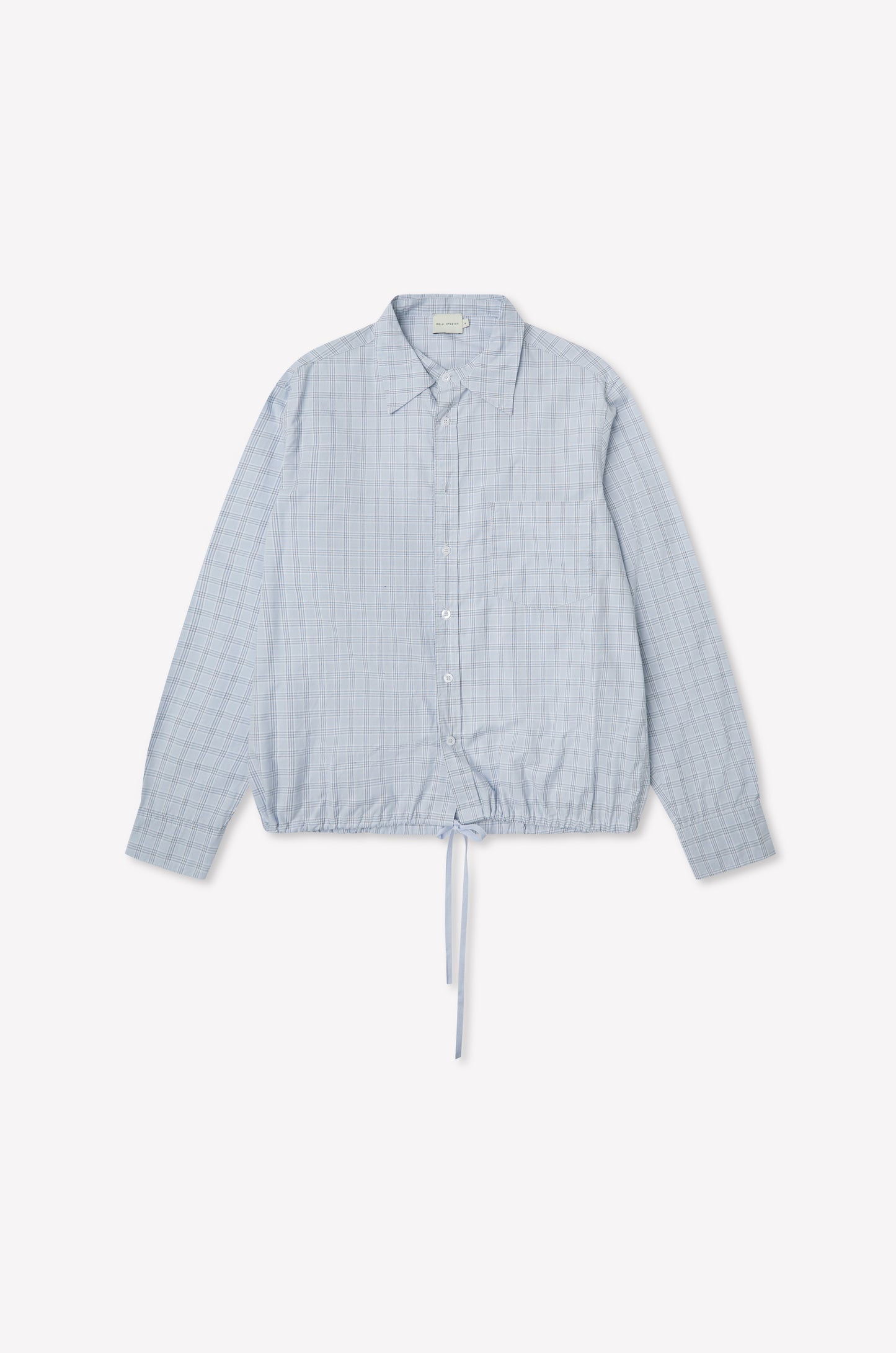 The Stand Shirt - Powder Check is a light blue oversized cotton shirt with long sleeves, subtle checkered pattern, chest pocket, classic collar, and drawstring hem. Shown on a plain white background, it channels deiji studios' relaxed style.