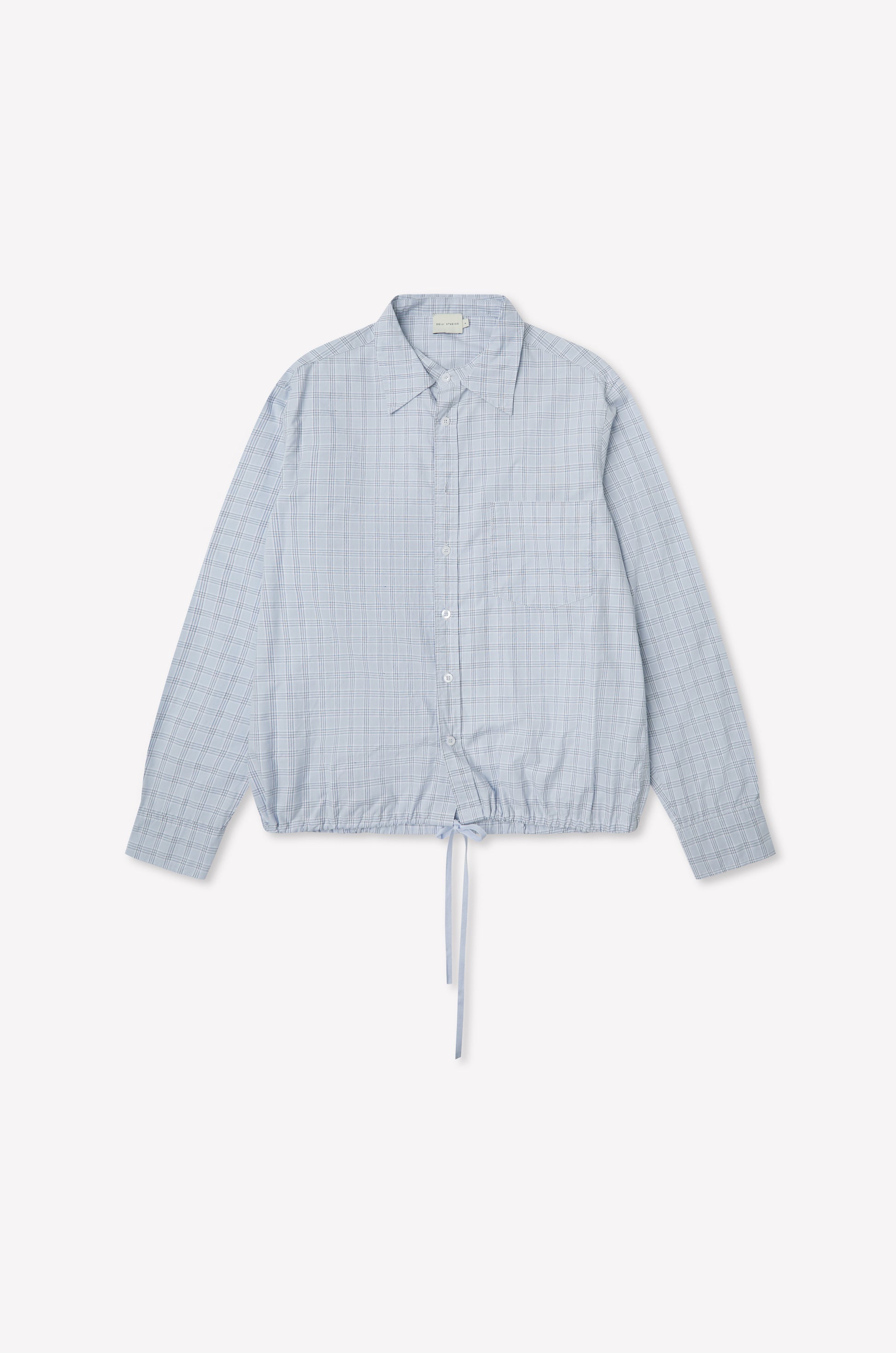 The Stand Shirt - Powder Check is a light blue oversized cotton shirt with long sleeves, subtle checkered pattern, chest pocket, classic collar, and drawstring hem. Shown on a plain white background, it channels deiji studios' relaxed style.