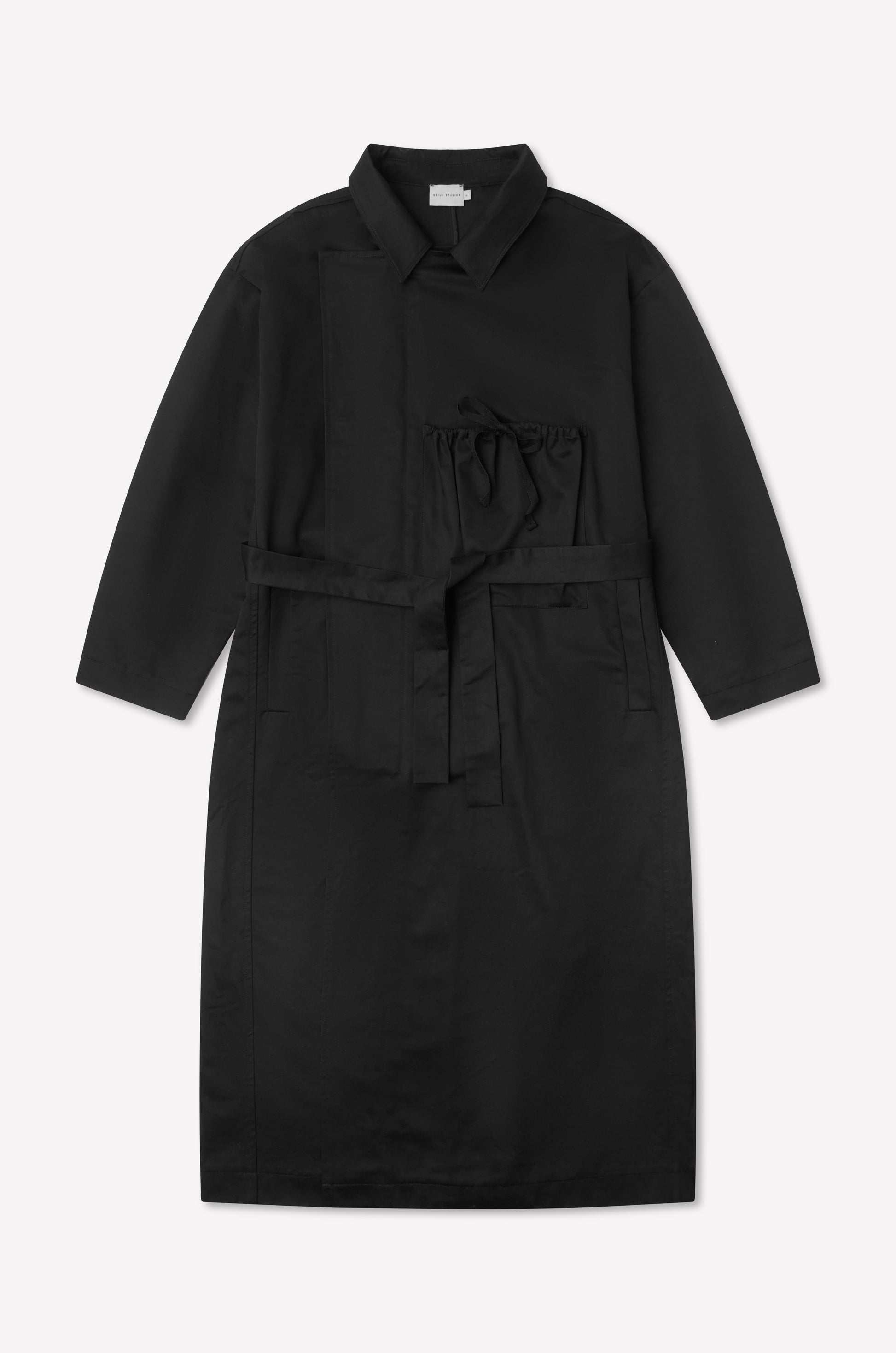 The Longline Jacket - Black from Deiji Studios is a long-sleeved, knee-length coat with a collar, front pocket, tie chest detail, and matching fabric belt, shown laid flat on a white background.