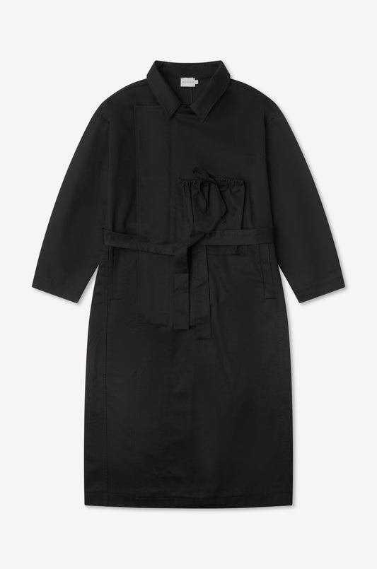 The Longline Jacket - Black from Deiji Studios is a long-sleeved, knee-length coat with a collar, front pocket, tie chest detail, and matching fabric belt, shown laid flat on a white background.