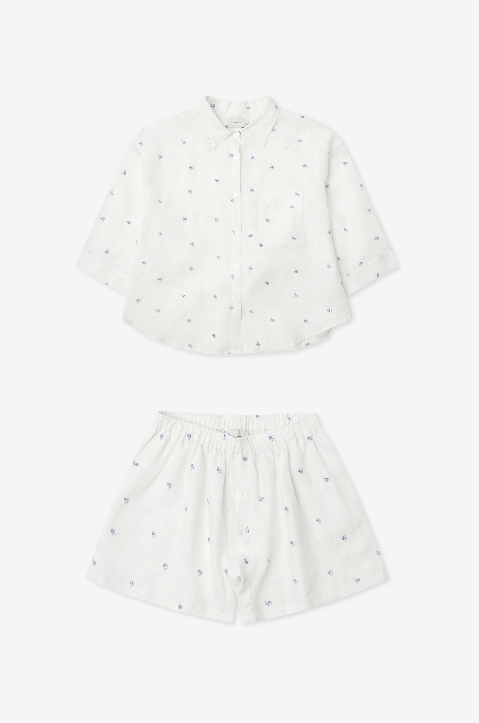 03 Set - Floral Blue: a children’s white outfit with long-sleeve oversized linen shirt and matching boxer-style shorts, both adorned with subtle blue floral embroidery. Shorts feature an elastic waistband. Displayed on a plain white background.