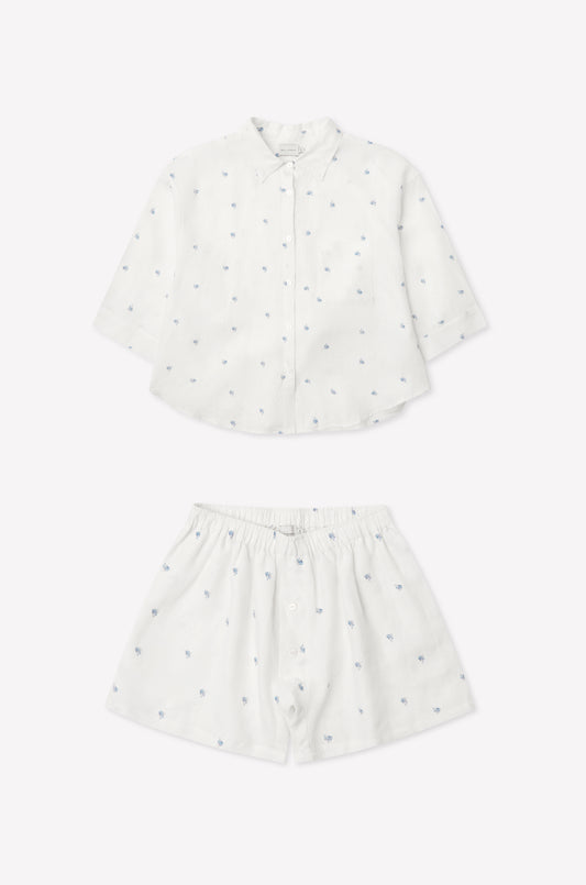03 Set - Floral Blue: a children’s white outfit with long-sleeve oversized linen shirt and matching boxer-style shorts, both adorned with subtle blue floral embroidery. Shorts feature an elastic waistband. Displayed on a plain white background.