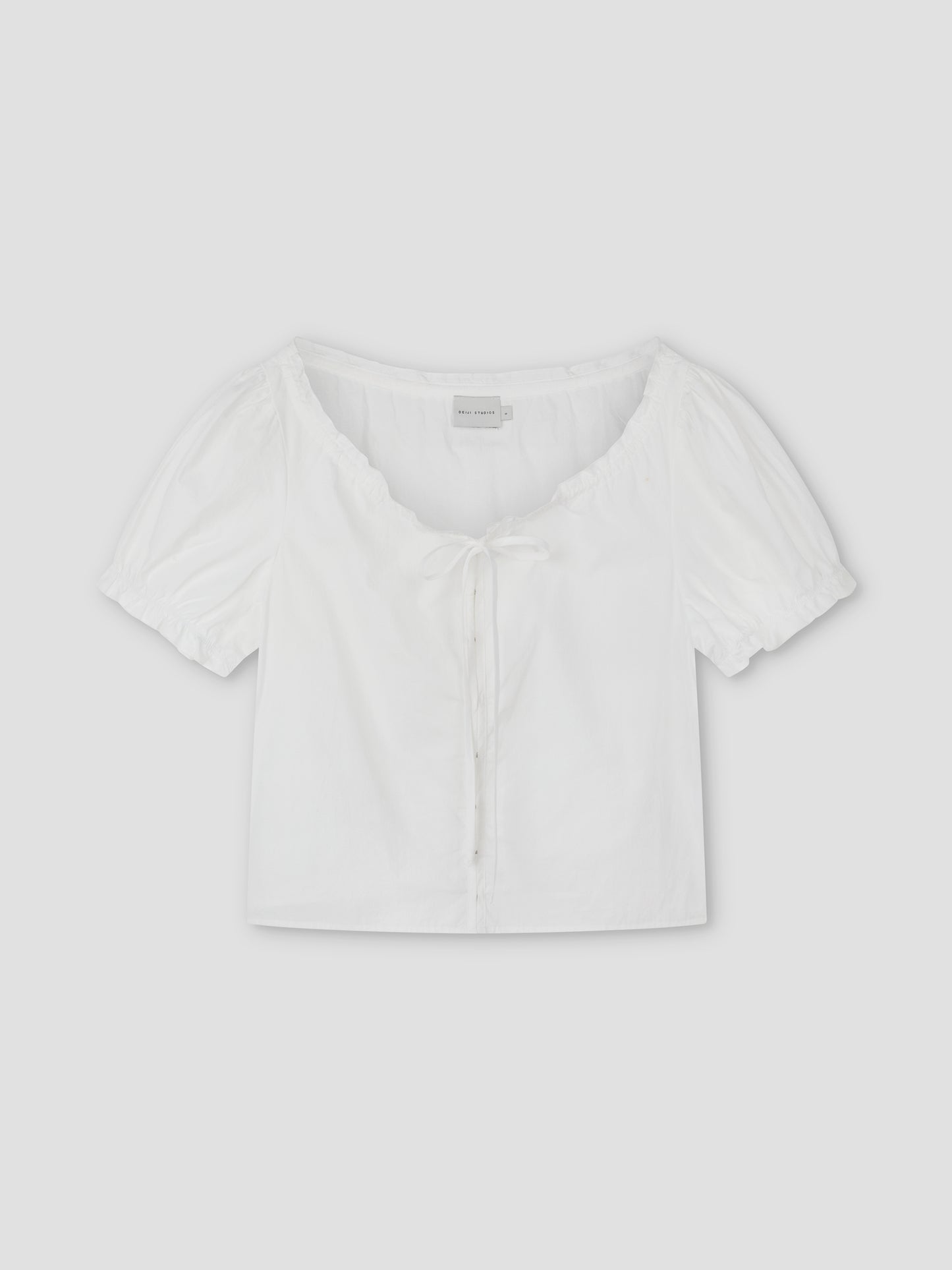 Close Shirt - White: A cropped short sleeve shirt with a scoop neckline, front tie detail, and gathered puff sleeves. Laid flat on a light gray background, this relaxed fit offers a stylish, comfortable option for casual looks.