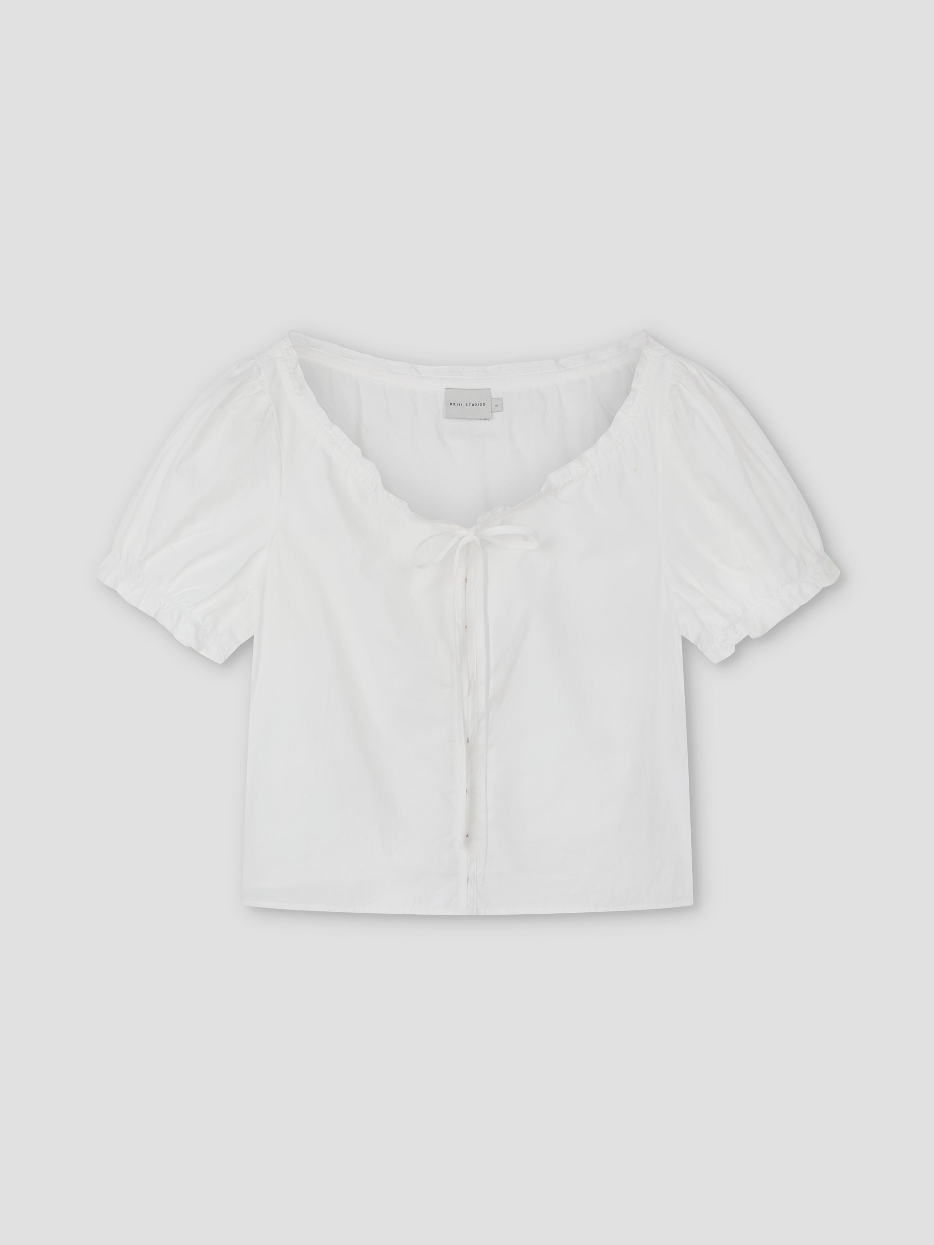 Close Shirt - White: A cropped short sleeve shirt with a scoop neckline, front tie detail, and gathered puff sleeves. Laid flat on a light gray background, this relaxed fit offers a stylish, comfortable option for casual looks.