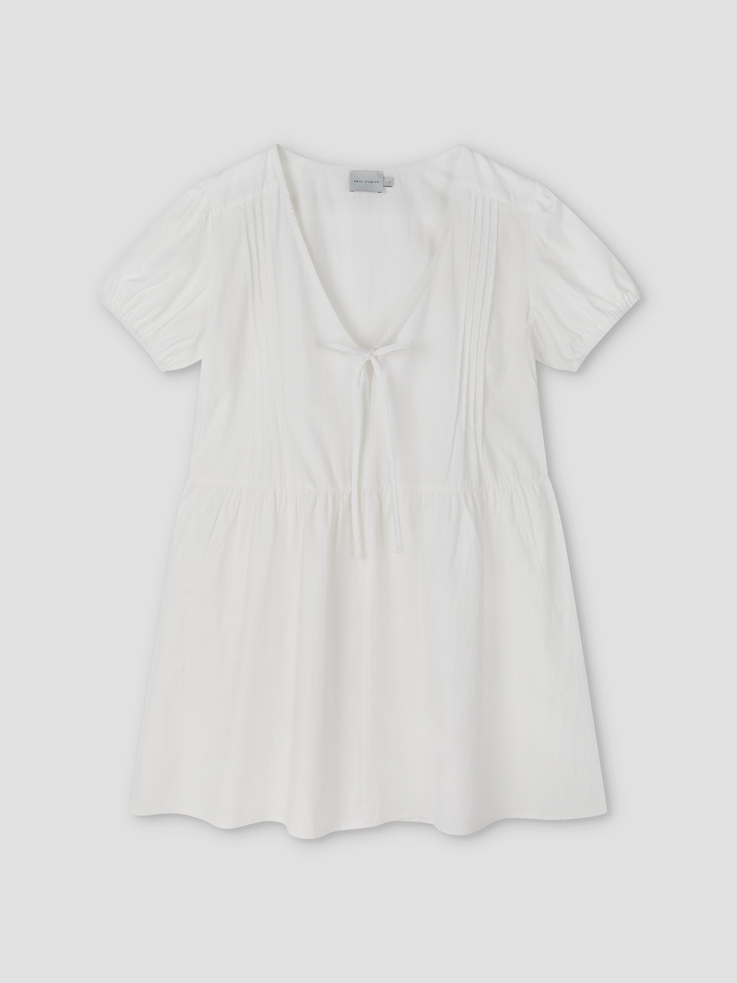 The V Pleat Dress - White Texture by Deiji Studios is a short-sleeve, lightweight white mini dress with a deep V-neckline, gathered waist, front tie detail, and subtle bodice pleats, shown laid flat on a light background.