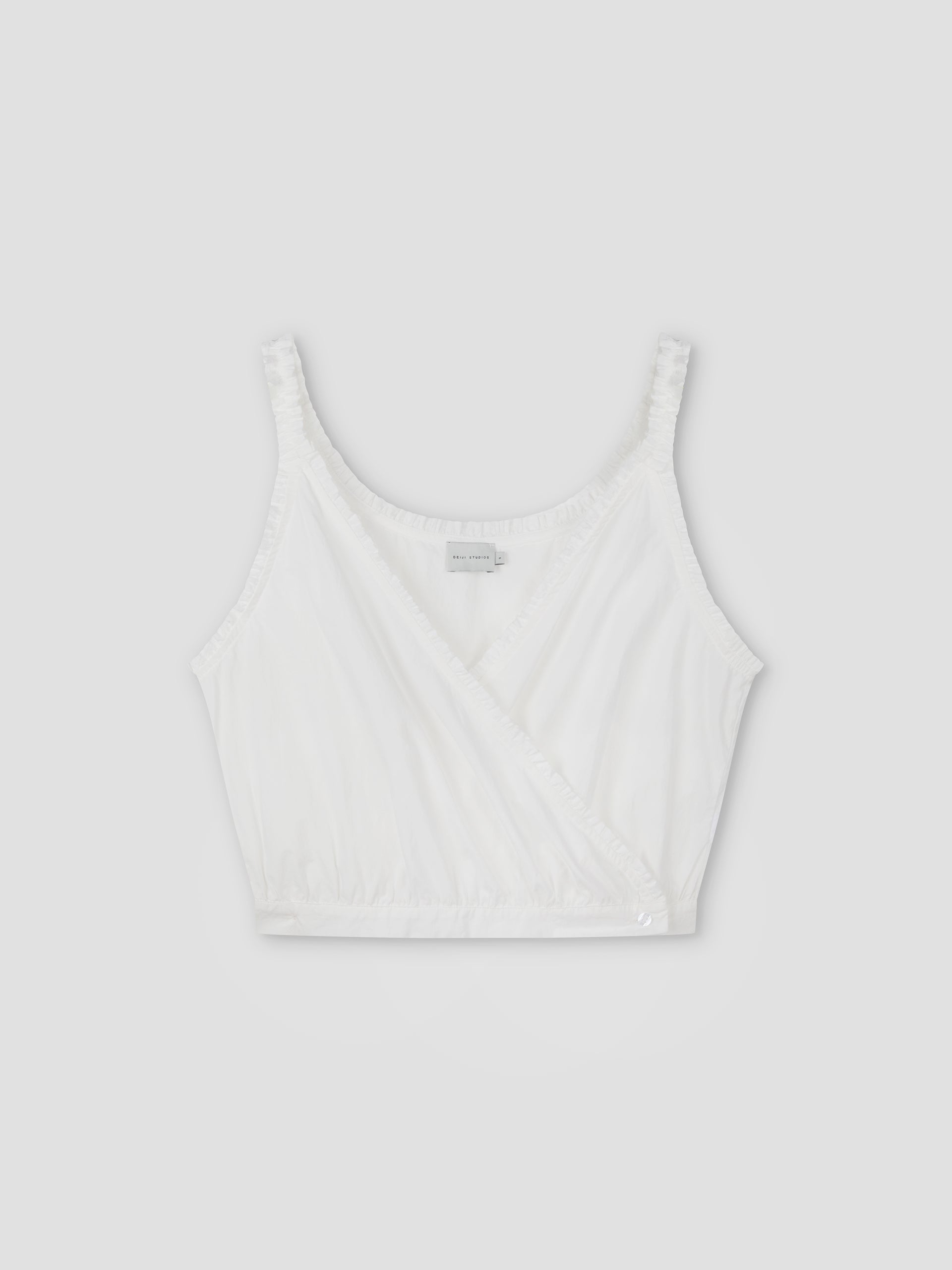 The Overlie Top - White features thin shoulder straps, a sleeveless wrap-style front, and gathered elastic at the hem, shown against a plain light gray background.