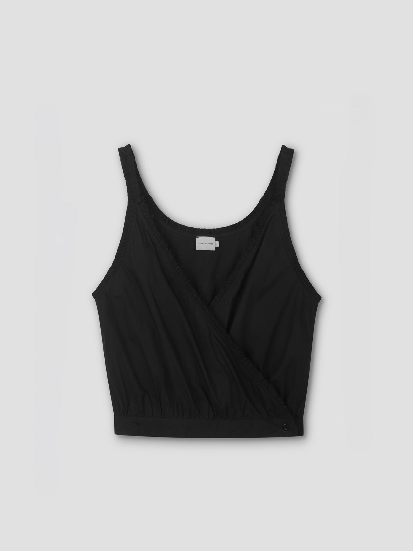 The Overlie Top - Black by Deiji Studios features a sleeveless, wrap-style front with a gathered hem and an interior label, shown against a light gray background.