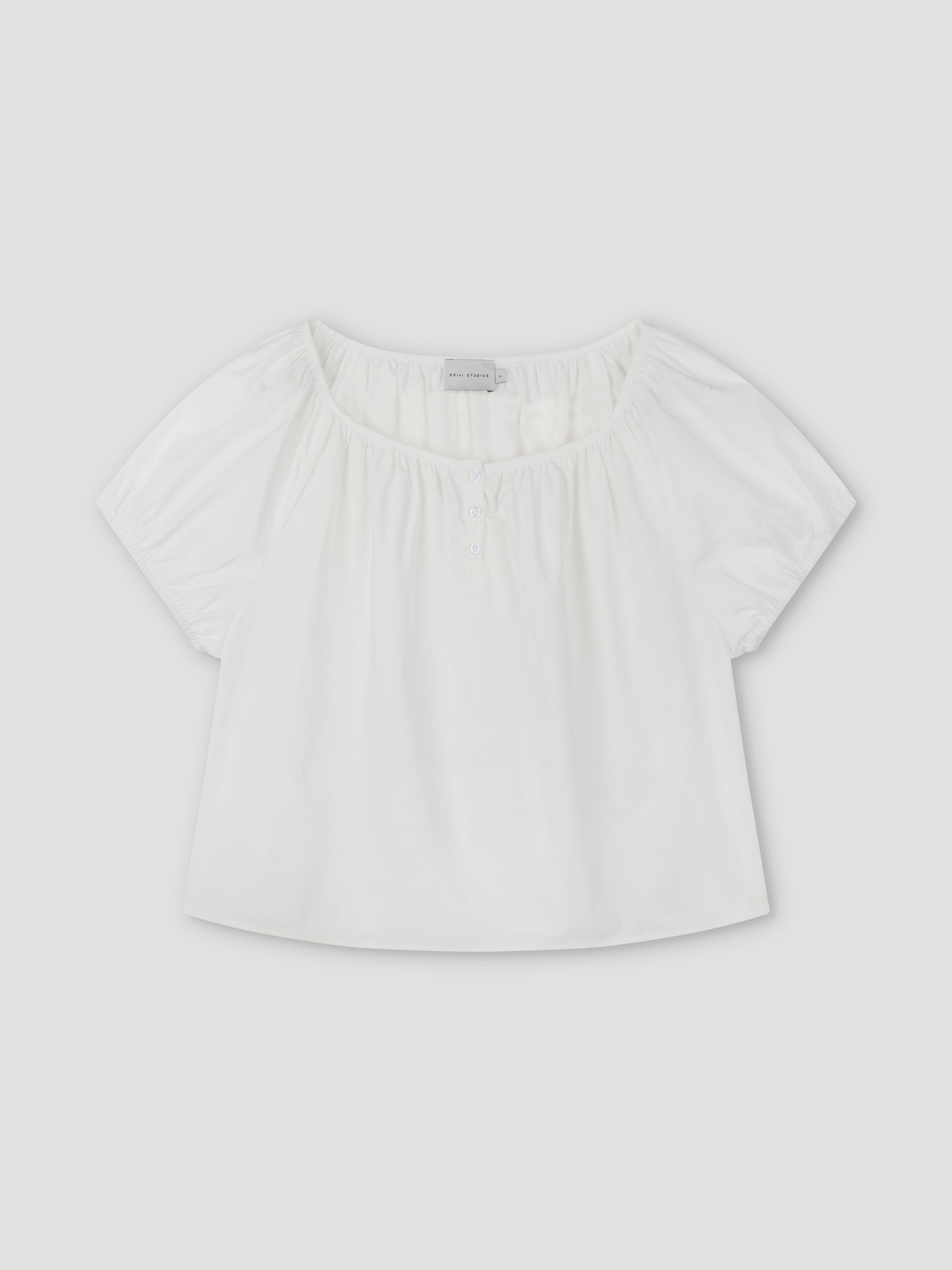 The Poplin T Shirt - White by Deiji Studios is a loose-fitting, short-sleeved blouse with puffed sleeves, a wide gathered neckline, and small front buttons, shown on a plain light gray background.