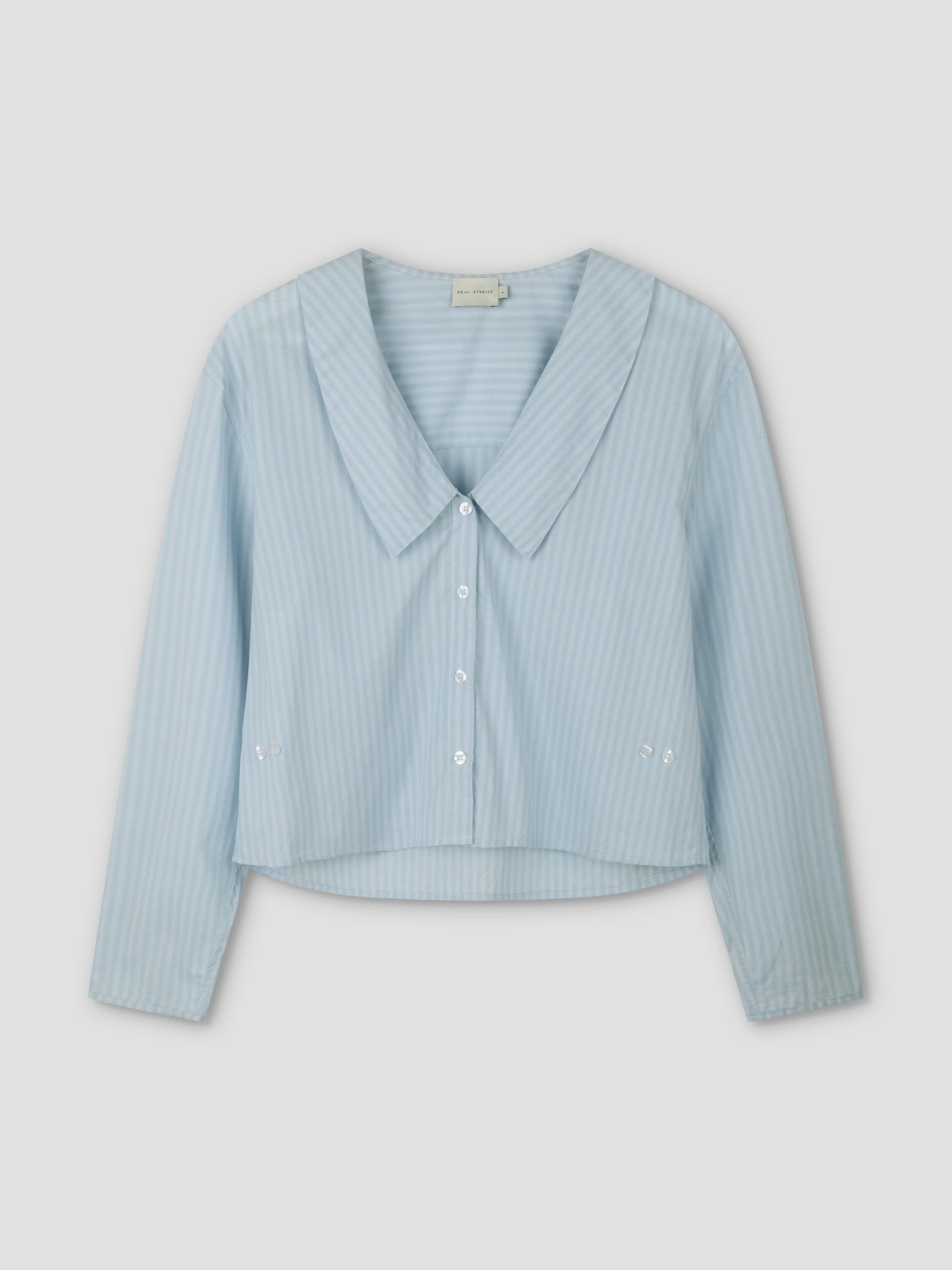 The Waist Button Shirt - Lake Blue Stripe features subtle vertical white stripes, a wide collar, cropped silhouette, long sleeves, and button-up front, displayed on a plain light background.