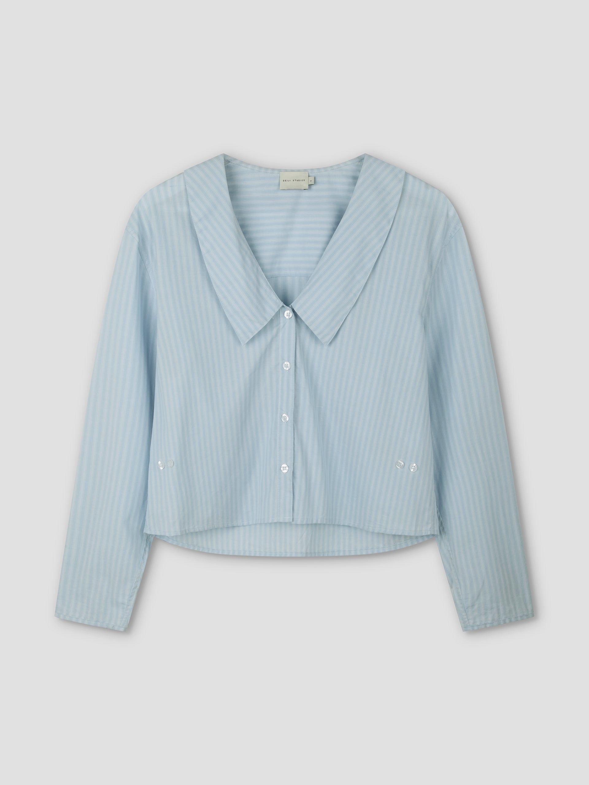 The Waist Button Shirt - Lake Blue Stripe features subtle vertical white stripes, a wide collar, cropped silhouette, long sleeves, and button-up front, displayed on a plain light background.