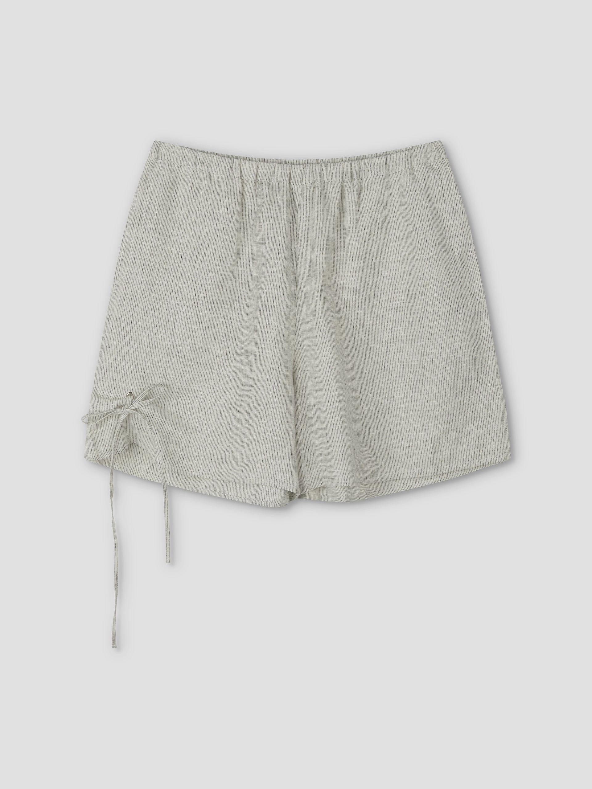 The Eyelet Short - Fine Stripe features a light gray linen, elastic waistband, and a decorative left tie, shown on a plain light background. Inspired by Deiji Studios' signature relaxed style.