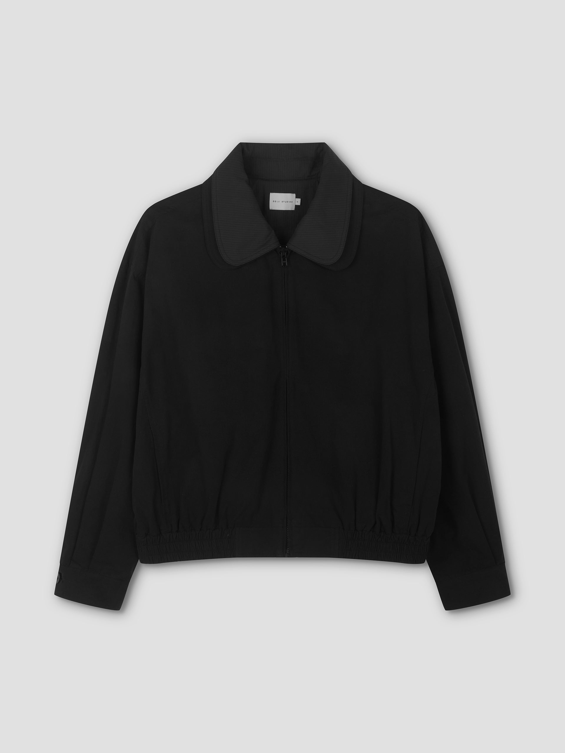 The Collared Zip Jacket - Black Contrast features long sleeves, a wide double-layered collar, and an elasticized hem, shown against a plain light grey background.