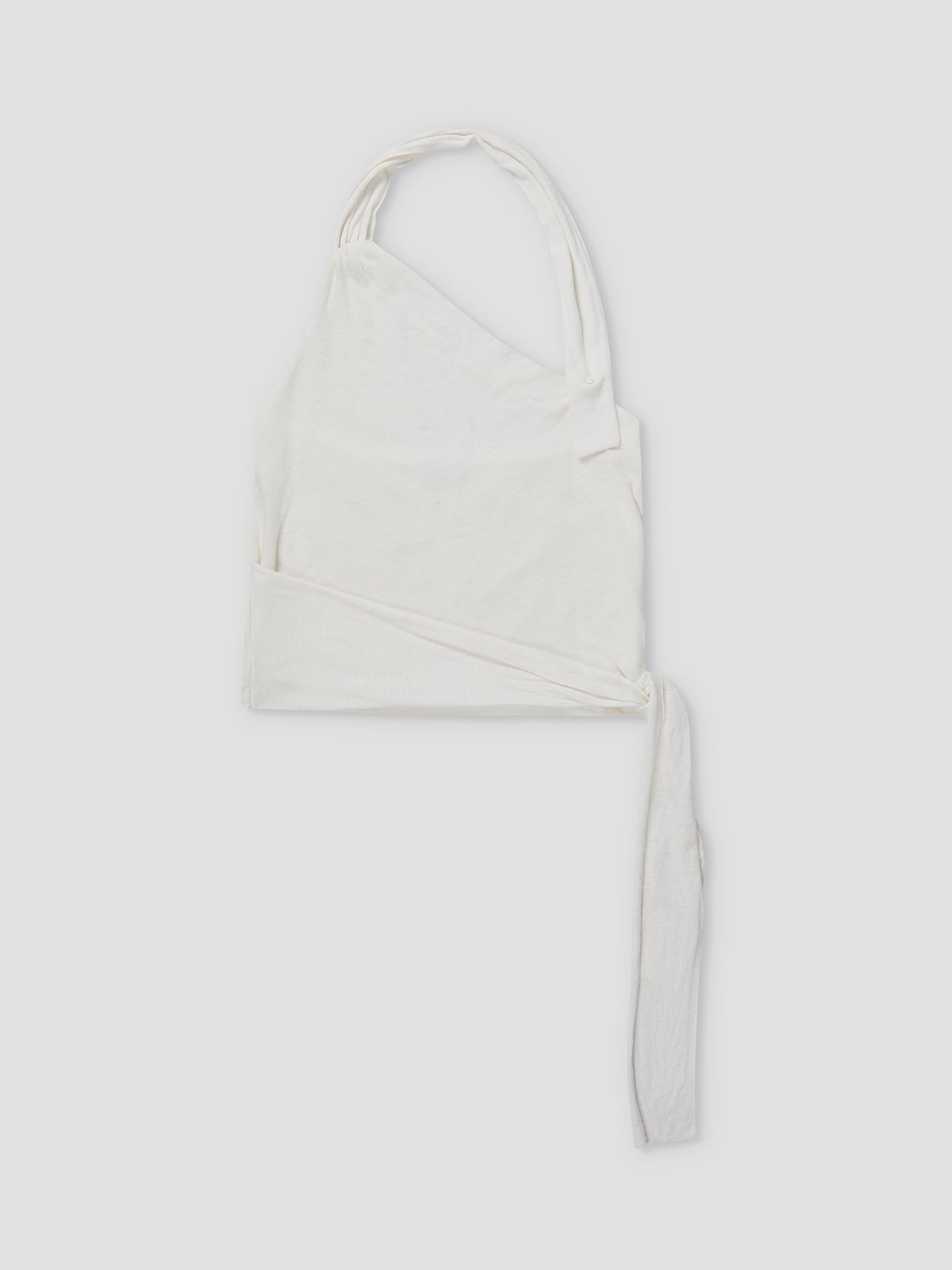 The Drape Halter - White is a white, asymmetrical one-shoulder crop top with a thick strap, long tie detail, and gathered neckline, shown laid flat on a light gray background.