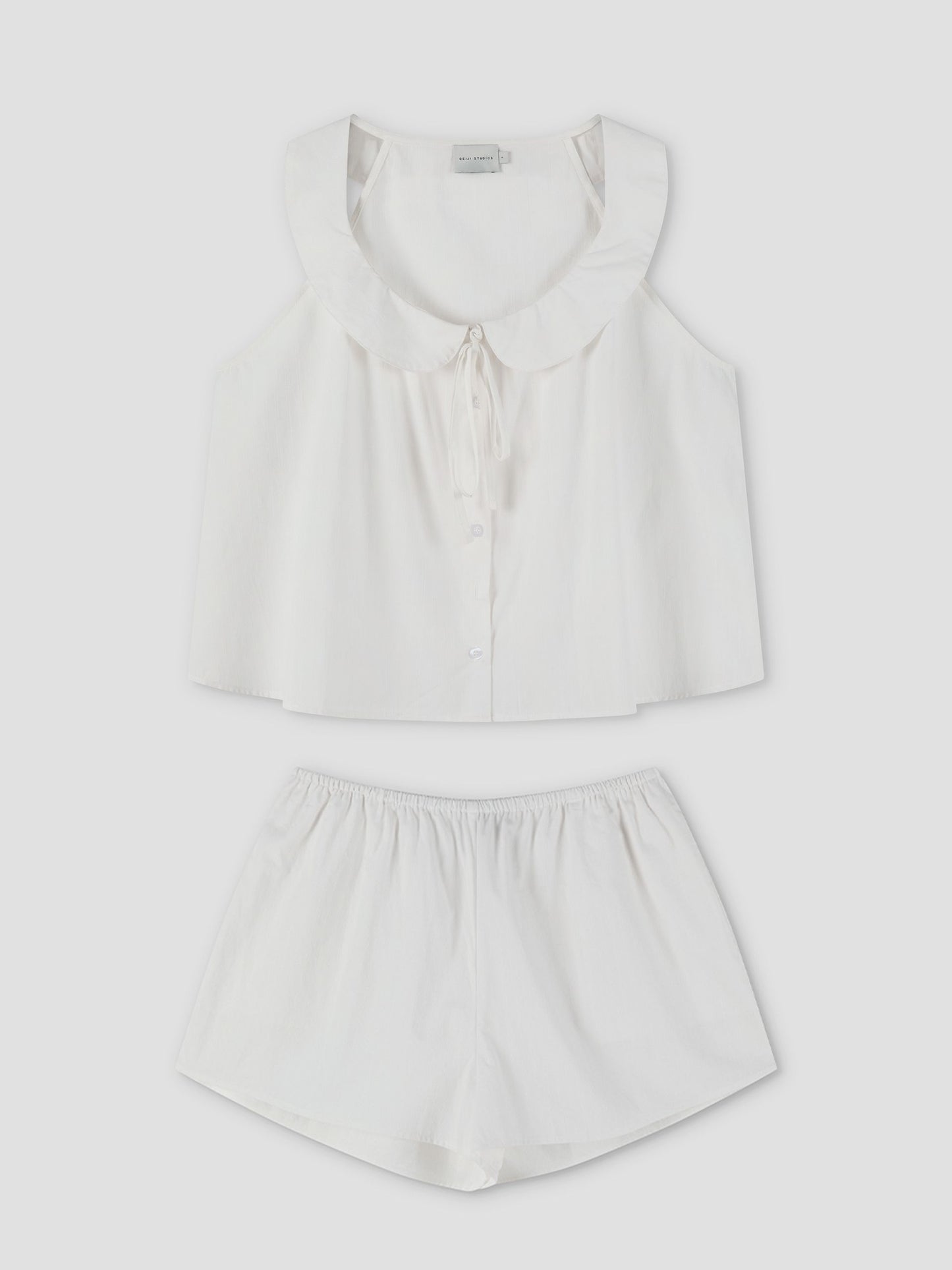 The Round Collar Set - White Texture features a white sleeveless pajama top with a ruffled collar and tie detail, button-up front, and matching loose-fitting shorts, displayed flat on a white background. Inspired by Deiji Studios.