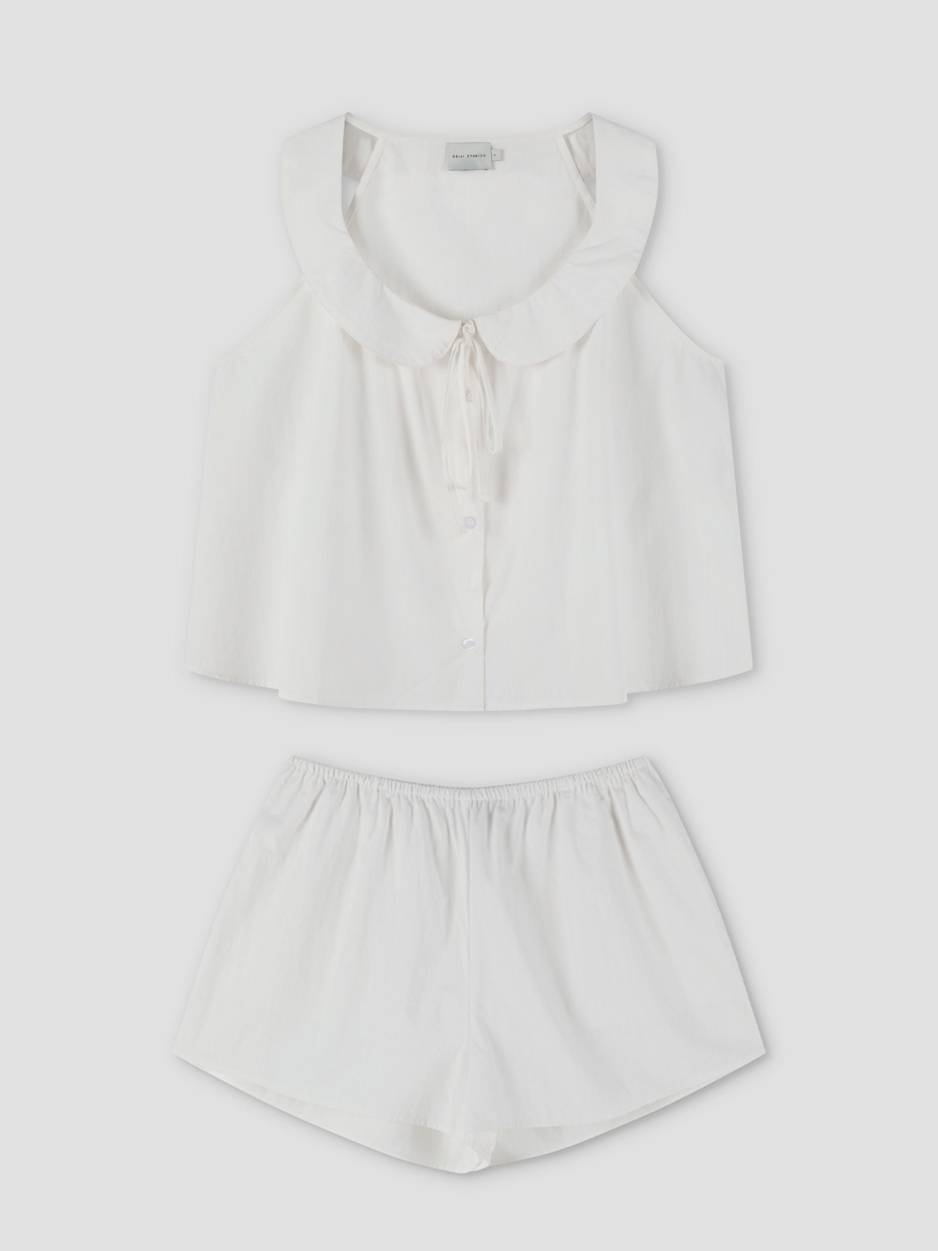 The Round Collar Set - White Texture features a white sleeveless pajama top with a ruffled collar and tie detail, button-up front, and matching loose-fitting shorts, displayed flat on a white background. Inspired by Deiji Studios.