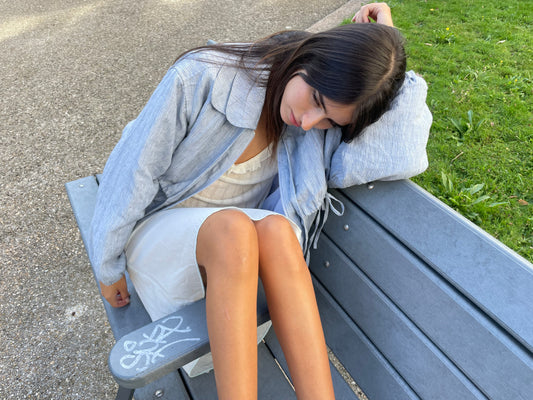 A young woman in a Double Layered Jacket - Blue Stripe and white dress sits on a gray bench, head resting on her arm, gazing down. Graffiti marks the bench’s armrest, with grass visible in the background.
